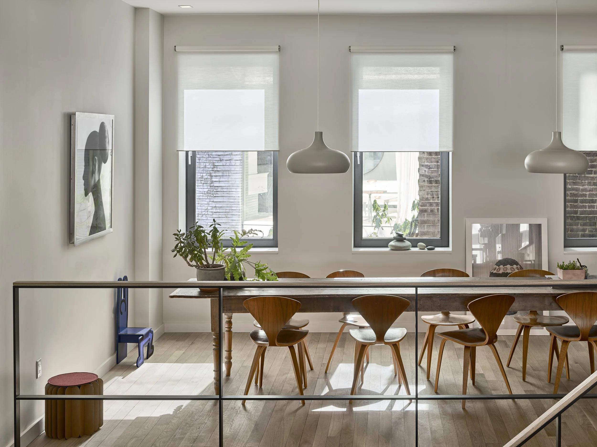 Interior of a modern dining area with wooden chairs around a wooden table, large windows with white blinds, and hanging pendant lamps, decorated with plants and framed artwork on white walls.