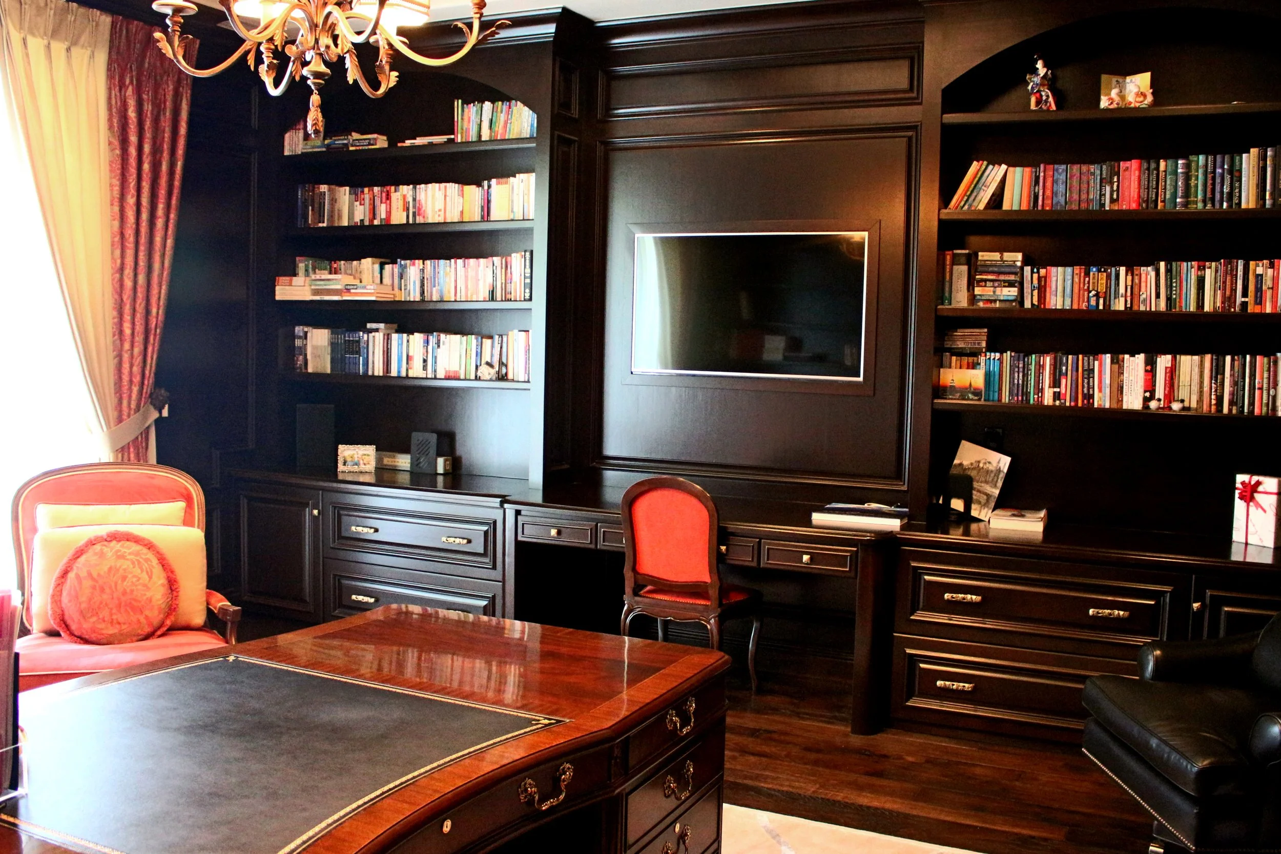 A room with dark wood shelves filled with books, a flat-screen TV, and a black desk, with red and gold furniture and a chandelier overhead.