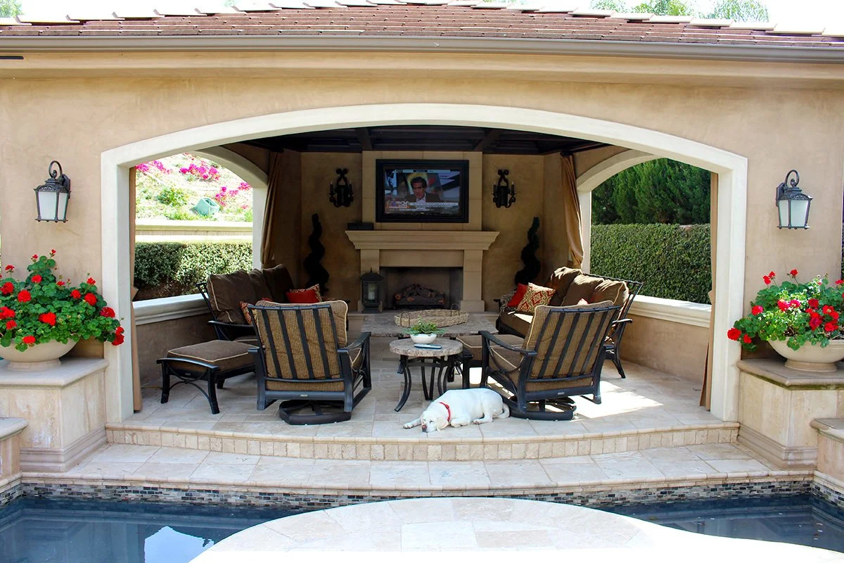 Outdoor patio with cushioned chairs, a small coffee table, a fireplace with a television mounted above, and a white dog lying on the tiled floor. Potted red flowers are on either side of the patio opening.