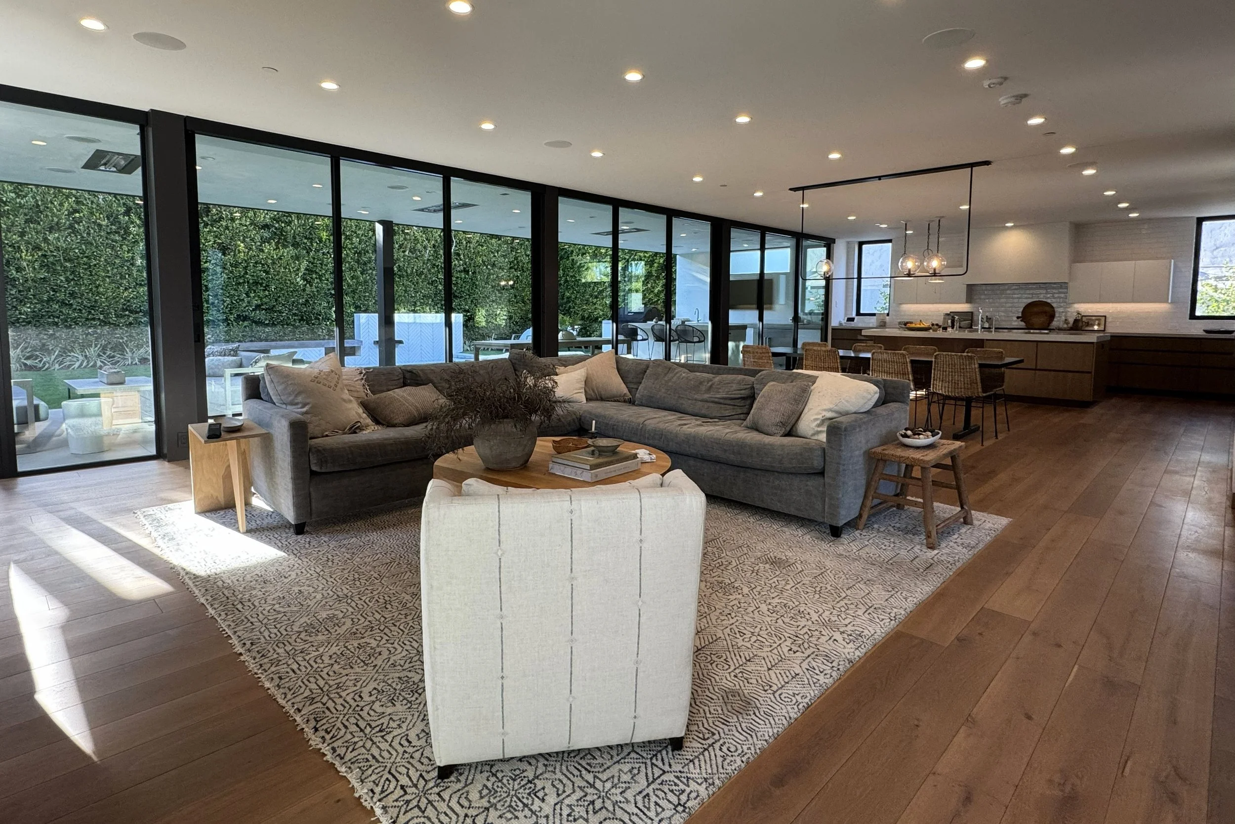 Modern open-concept living room and kitchen with large glass windows, a gray sectional sofa, white armchair, wooden coffee table, and dining area with bar stools, illuminated by ceiling lights.