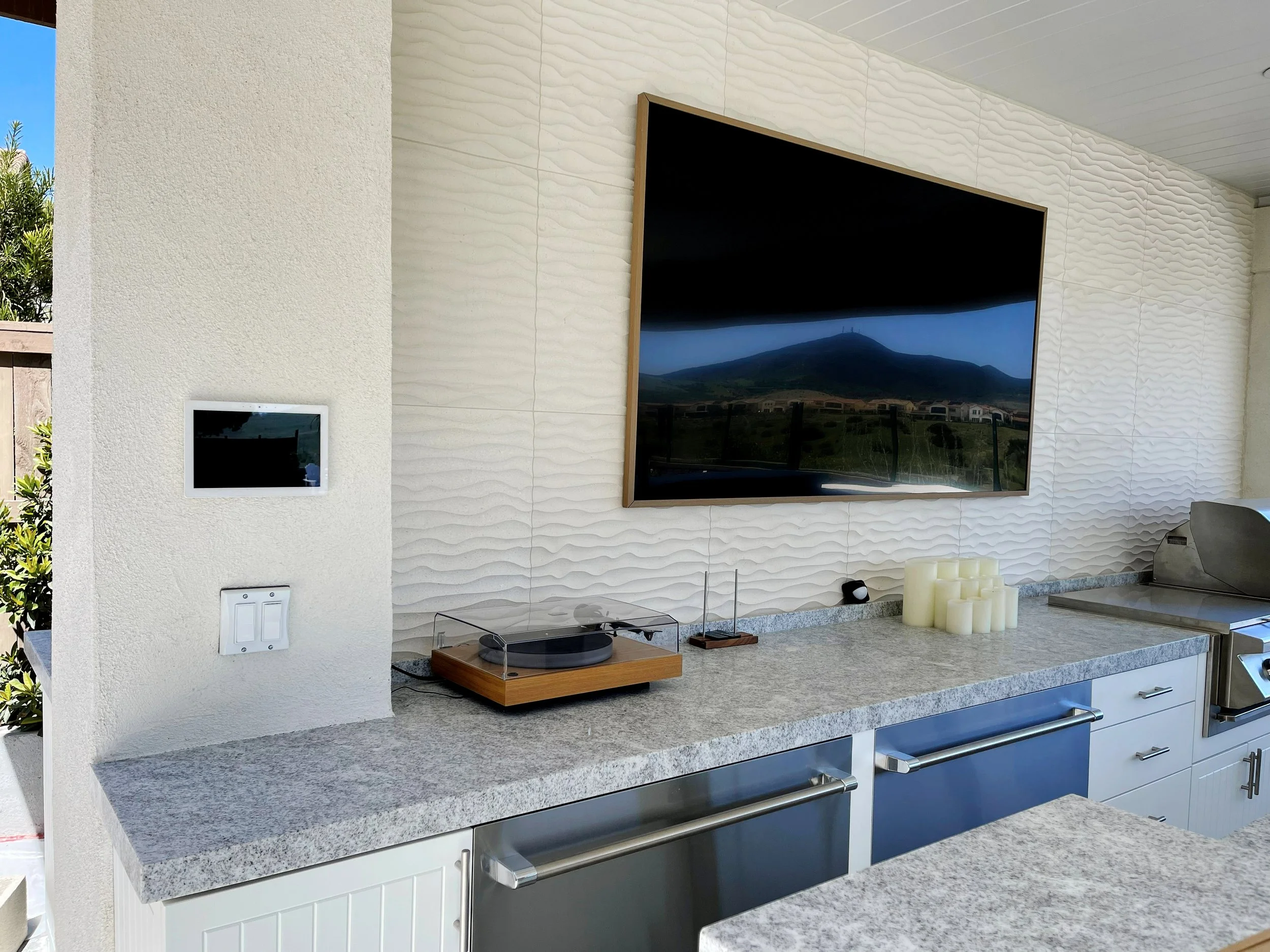 Outdoor kitchen with a granite countertop, a large flat-screen TV mounted on textured white wall, and a countertop speaker system, with a view of a mountain and blue sky reflected in the TV.