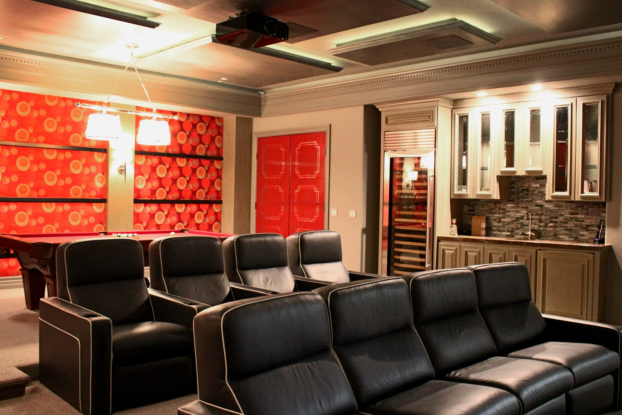 Interior of a living room with black leather chairs arranged in rows, a pool table with black cushions, a patterned red wall with pendant lights, a wine cooler, and built-in cabinets with a tiled backsplash in the kitchen area.