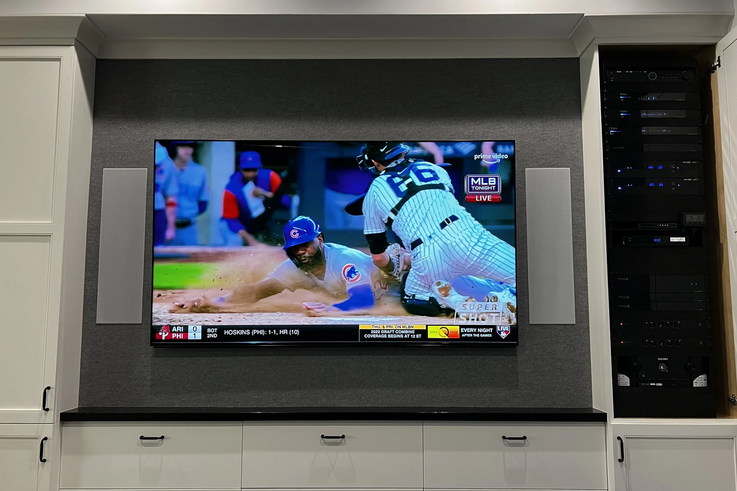 A television mounted on a white cabinet showing a baseball game, with two players involved in a play at home plate.