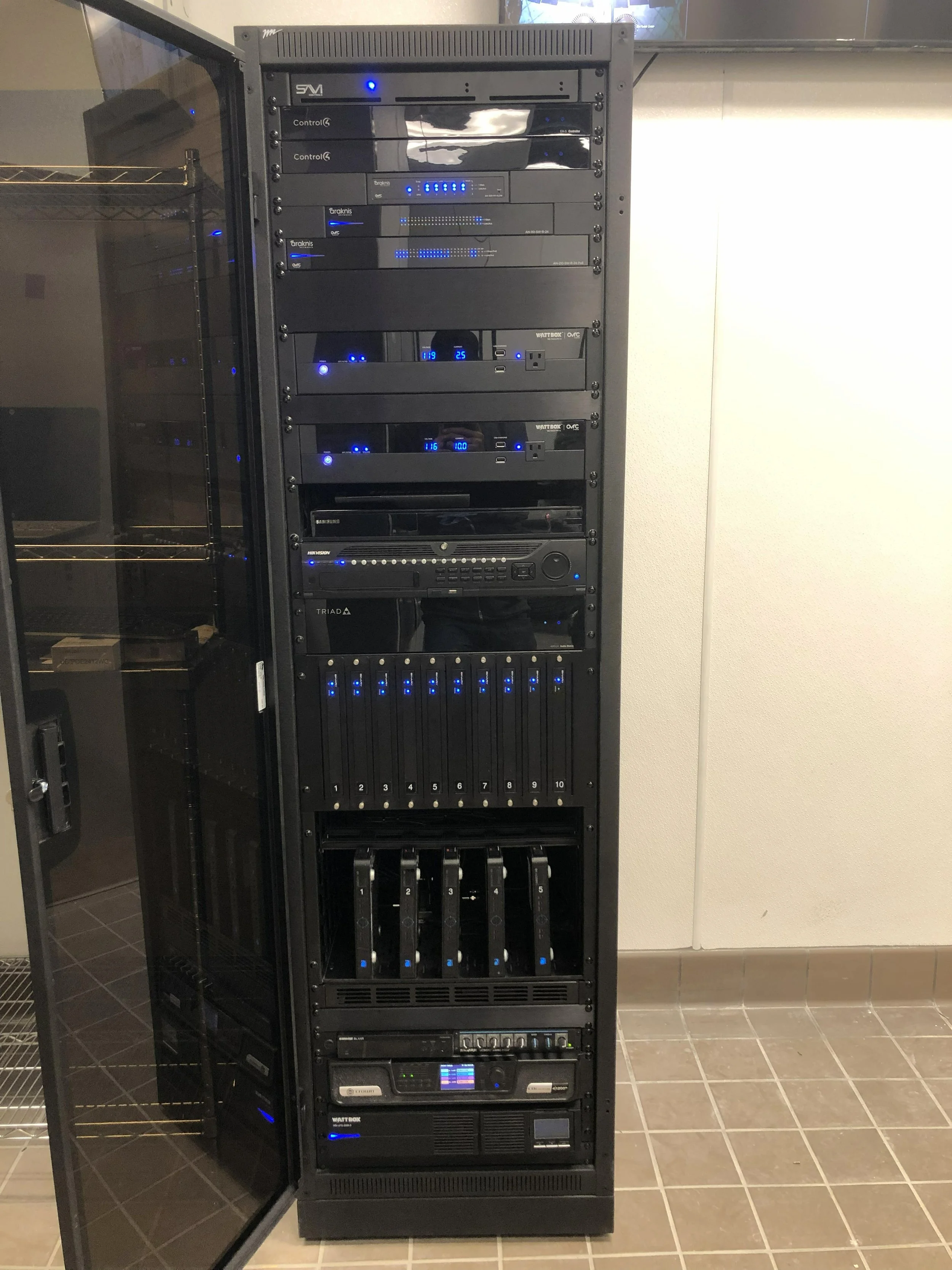 An open server rack filled with various electronic equipment, including switches, routers, and power supplies, located in a server room with tiled flooring and a wall in the background.