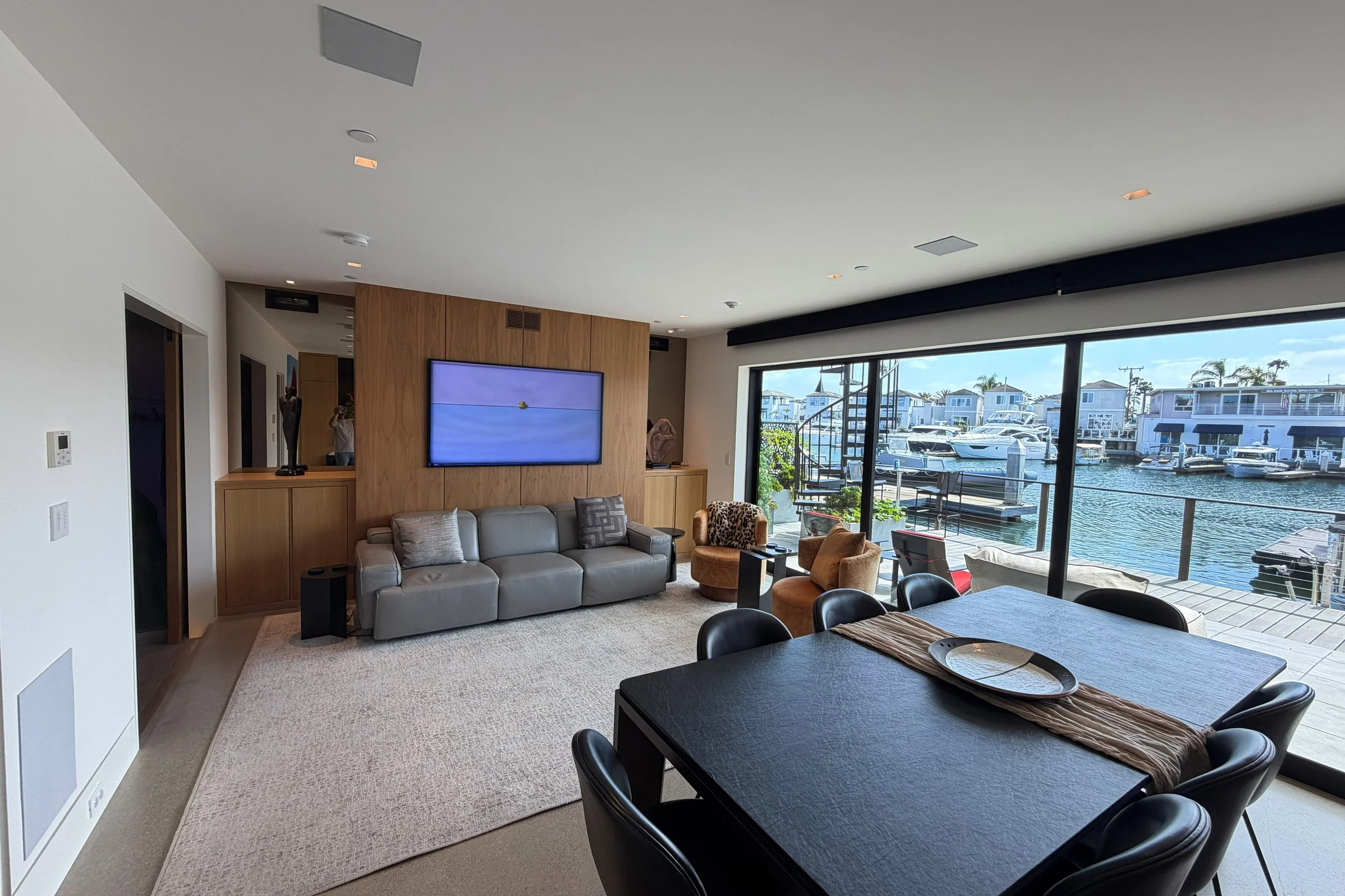 Living room with large glass sliding doors overlooking a marina with boats, including a sofa, chairs, a wall-mounted TV, and a dining table.