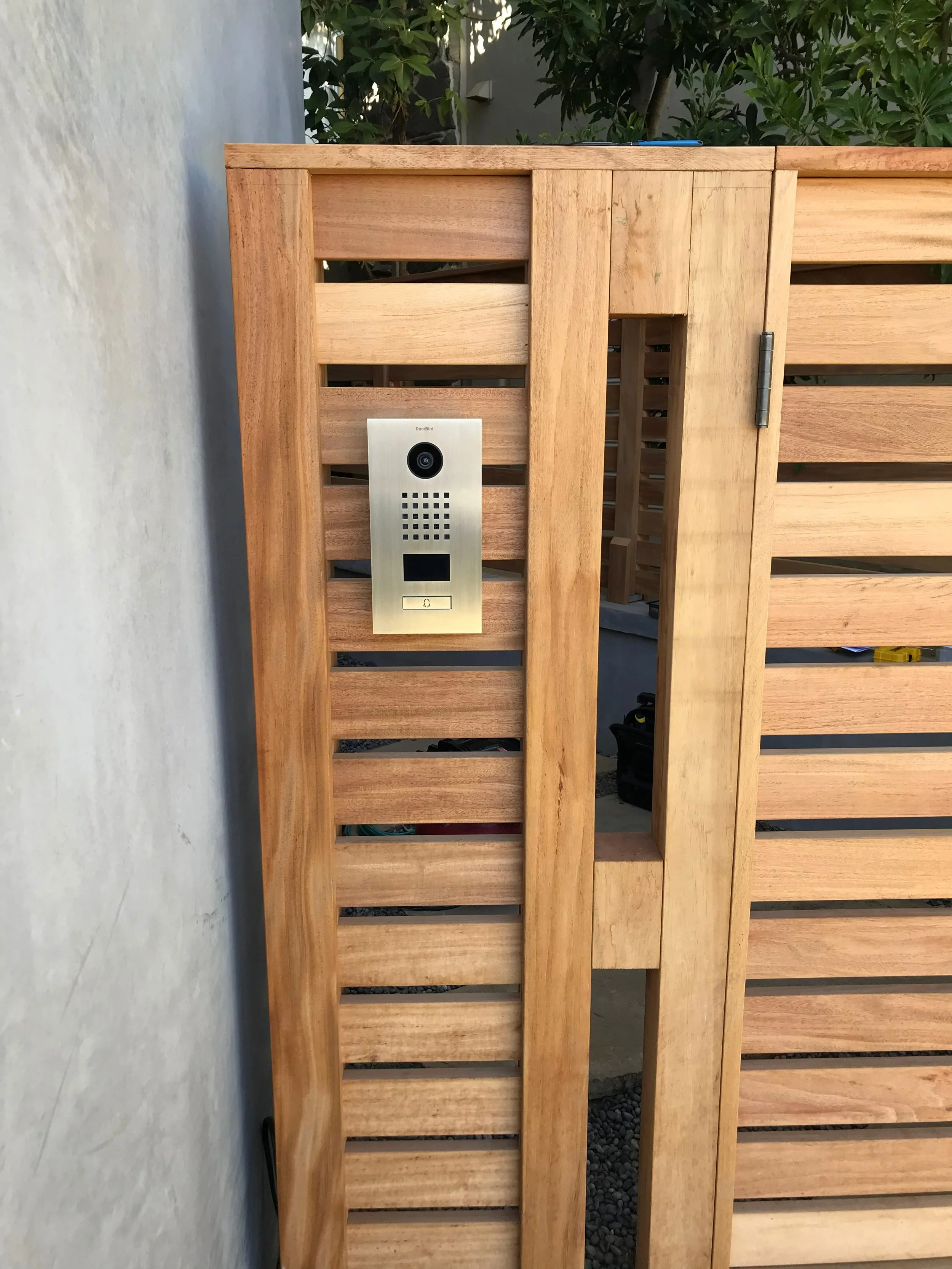 Wooden gate with a metal intercom and camera system installed in it, outdoors with greenery in the background.