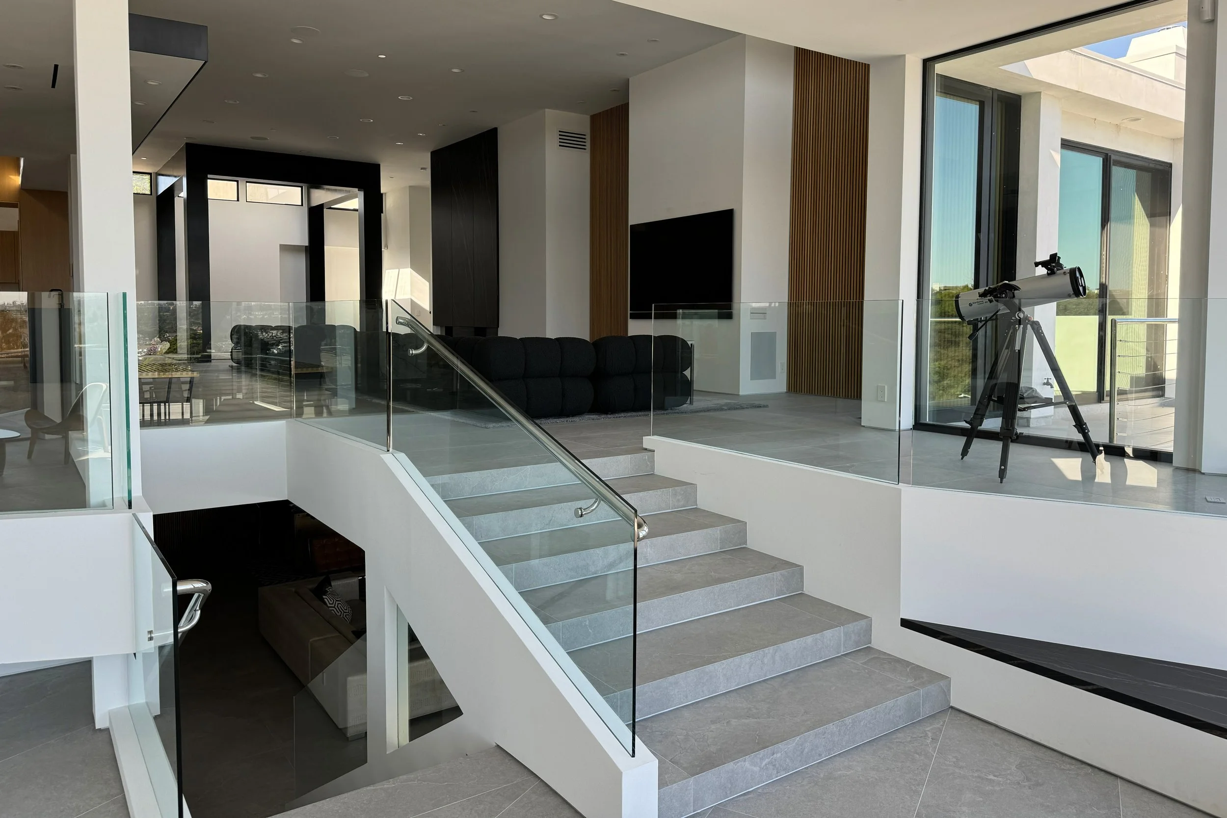 Modern interior living space with staircase, black sofa, television, telescope near large window, and views of outdoor greenery.