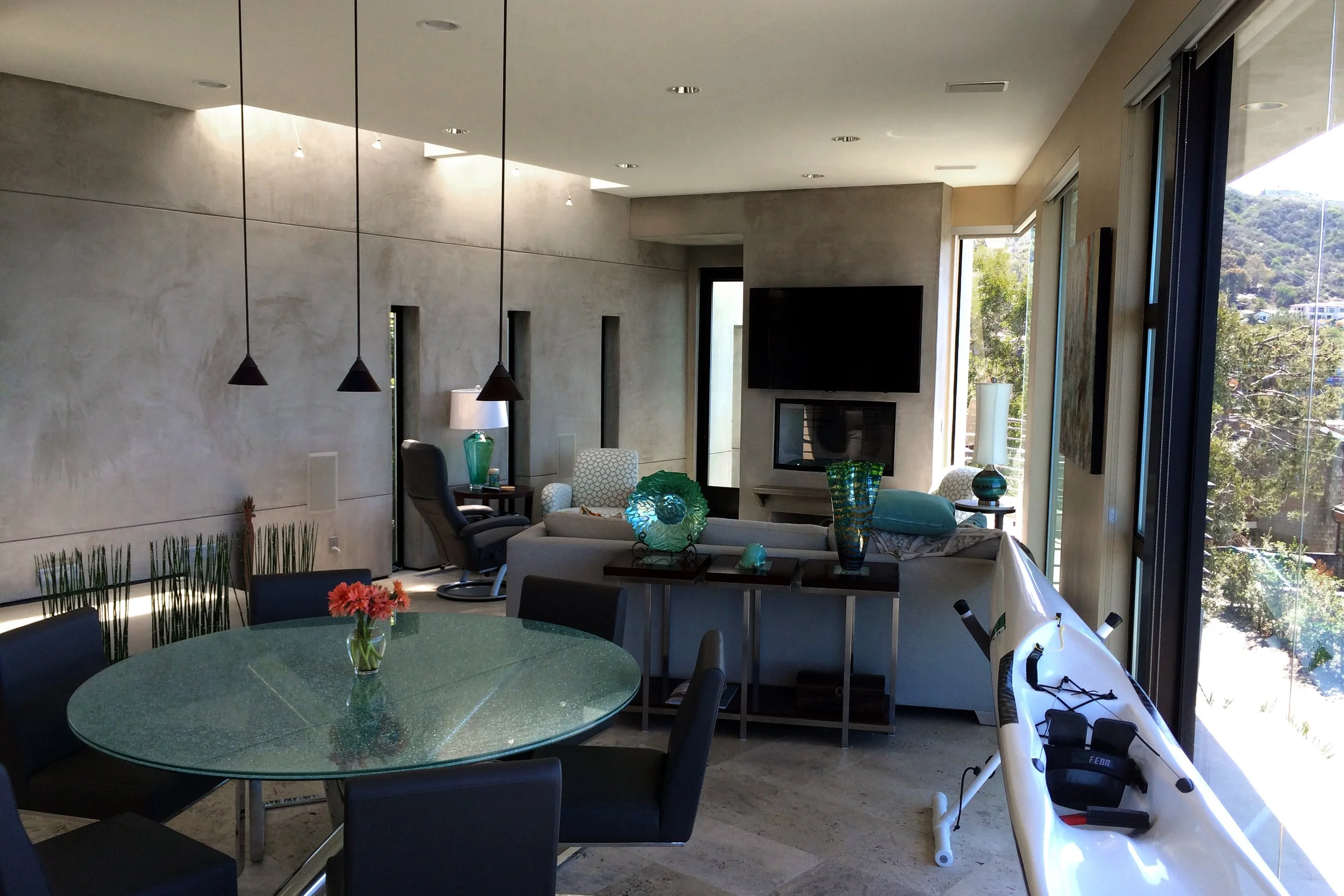 The image shows a modern living room with large windows, a flat-screen TV, and a seating area with chairs and a sofa adorned with decorative pillows. There is a round dining table with a flower arrangement in the foreground. The room has a minimalist