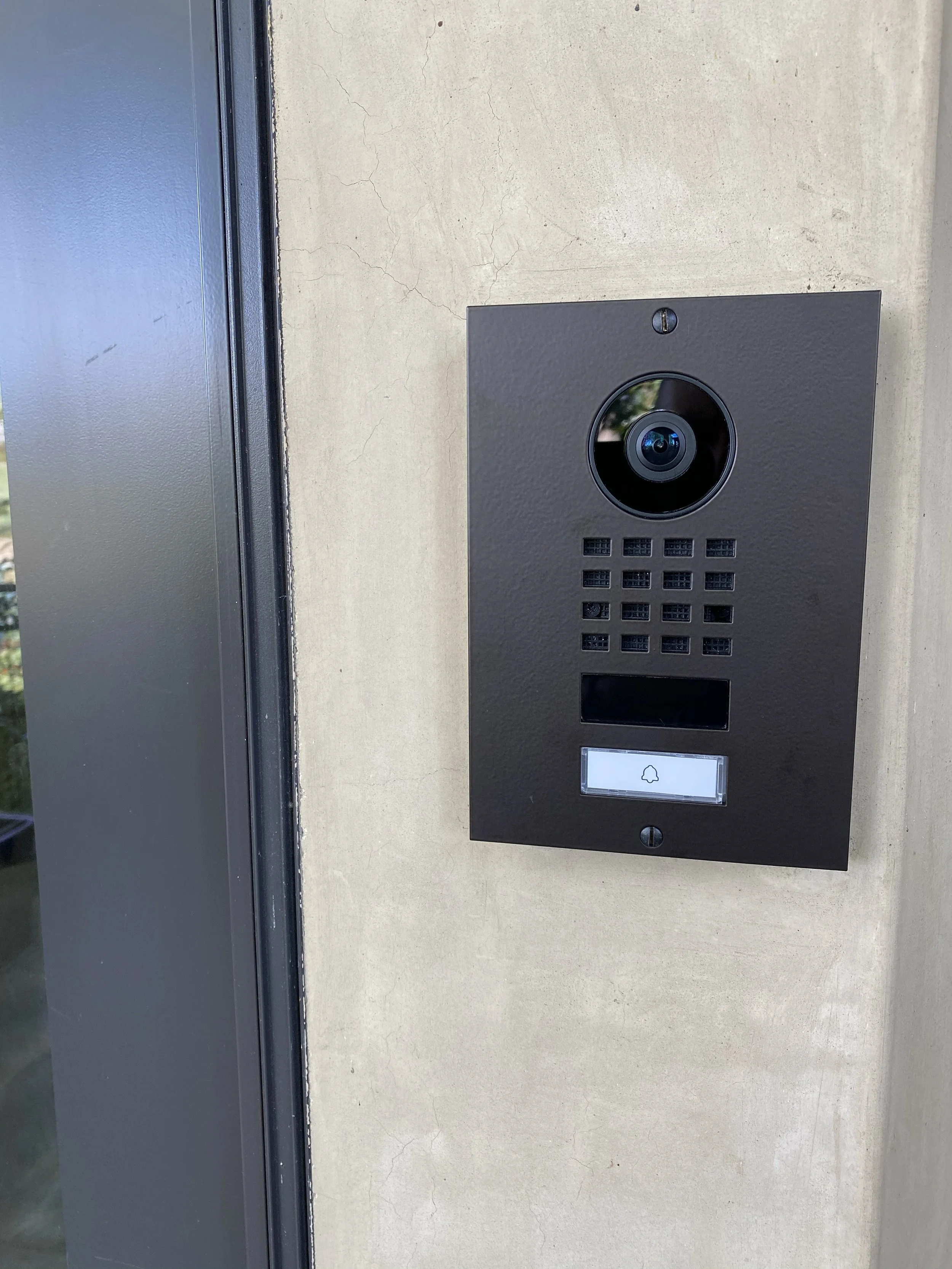 An intercom or doorbell system with a camera embedded in a black panel, mounted on a beige wall next to a gray door frame.