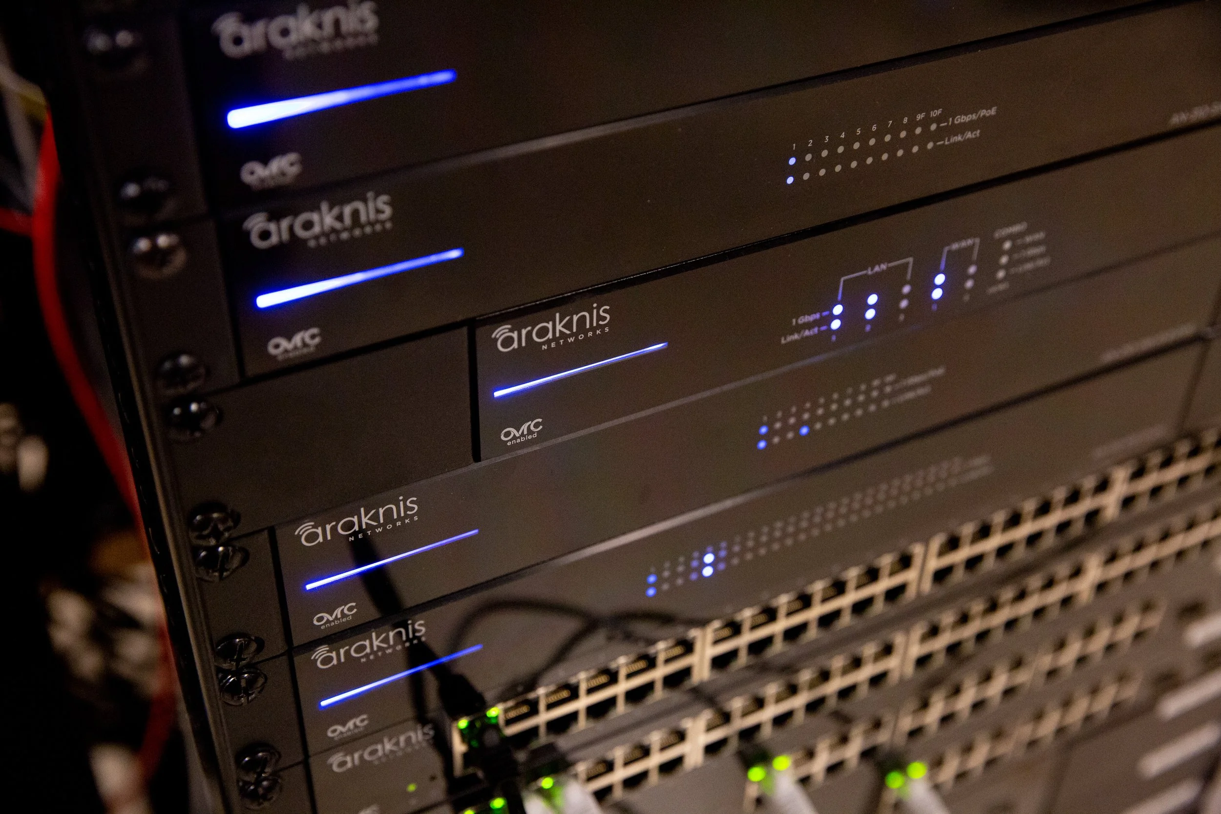 A stack of black Araknis Networks network switches with blue and green LED indicator lights, mounted in a server rack.