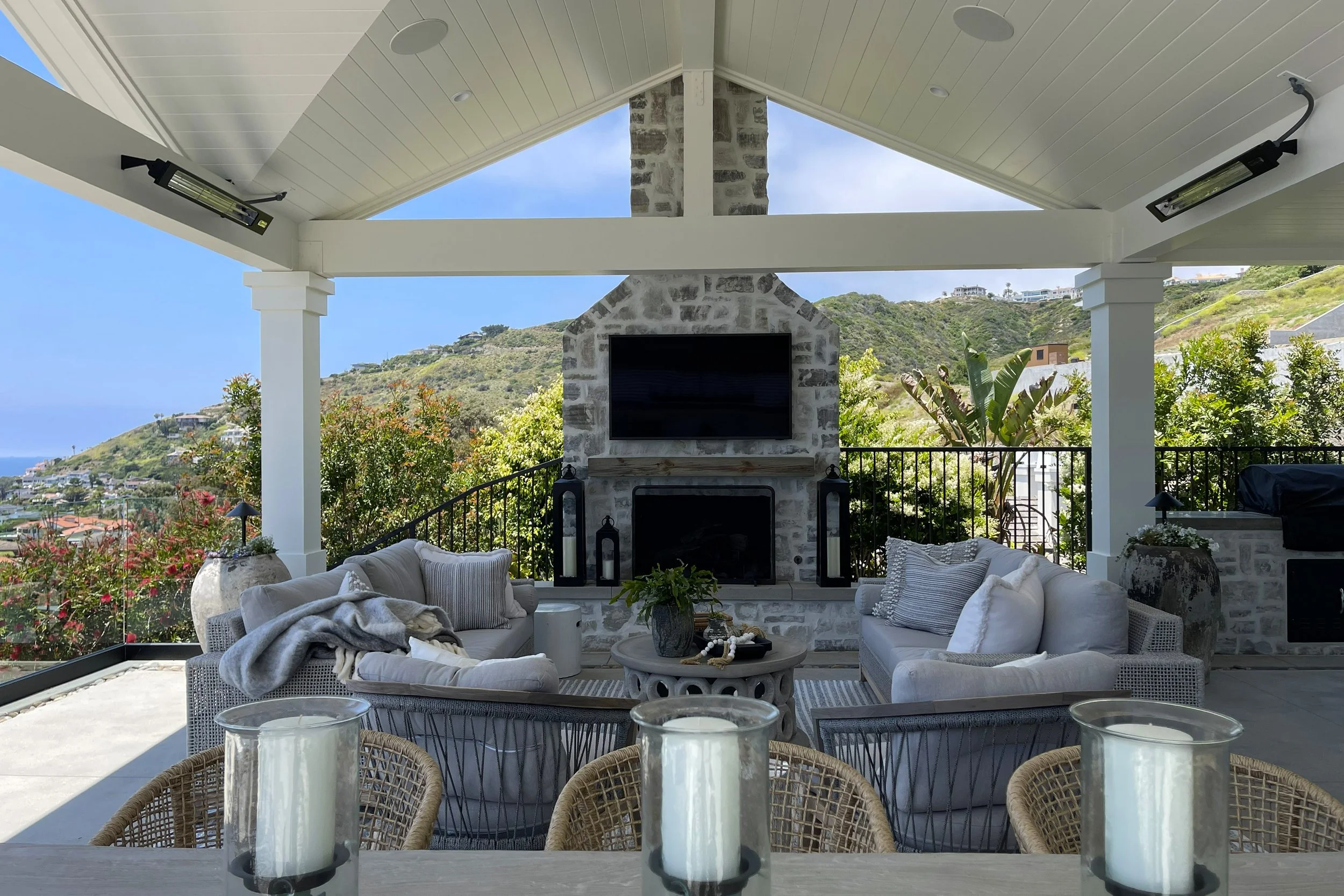An outdoor living space with cushioned seating, a stone fireplace with a mounted television, and a scenic hillside view in the background.