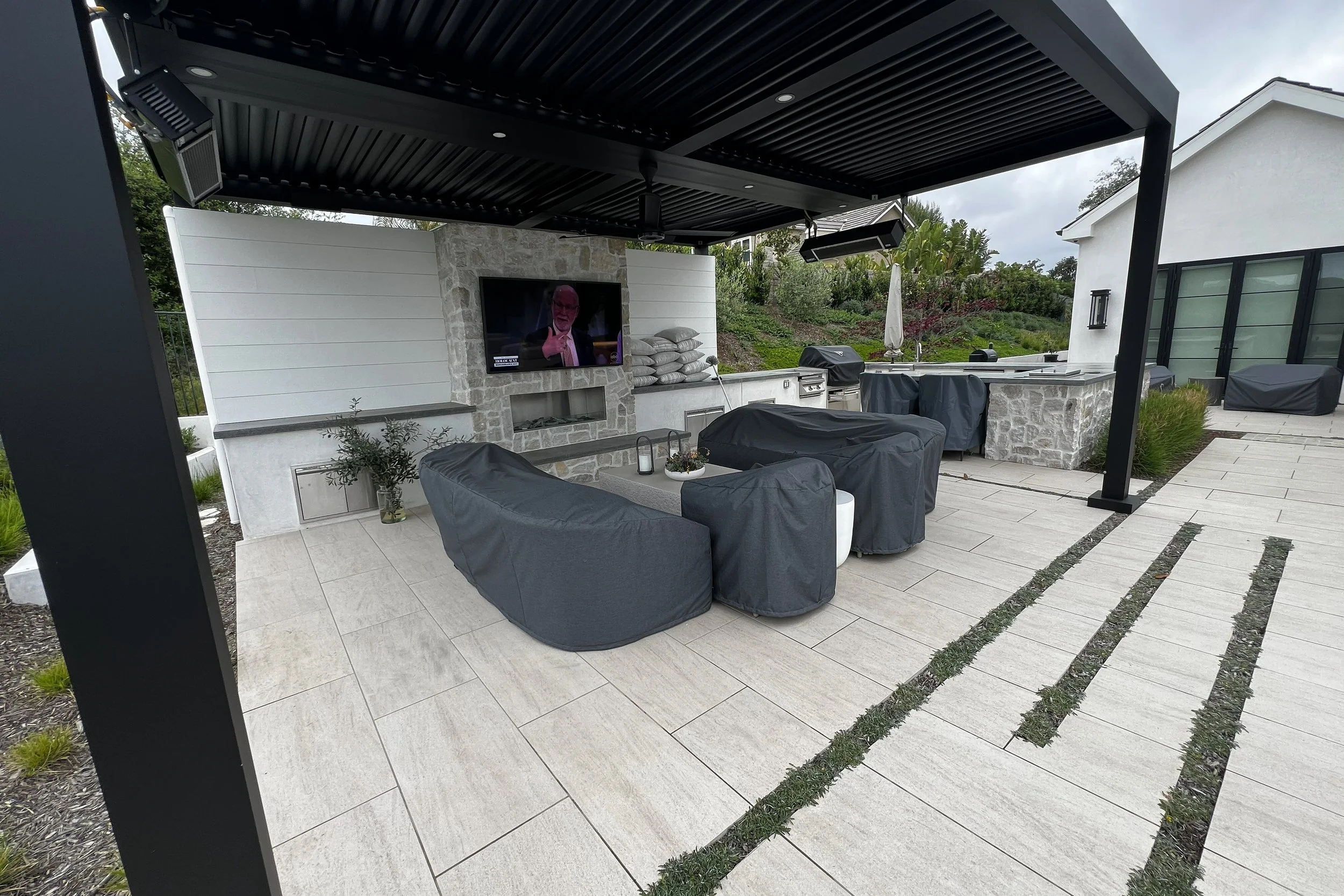 An outdoor patio with a ceiling, ceiling fans, and mounted speakers. There is a wall-mounted TV above a stone fireplace, with pillows stacked nearby. The patio has seating with covered chairs, and an outdoor kitchen area with a sink and grill. The fl