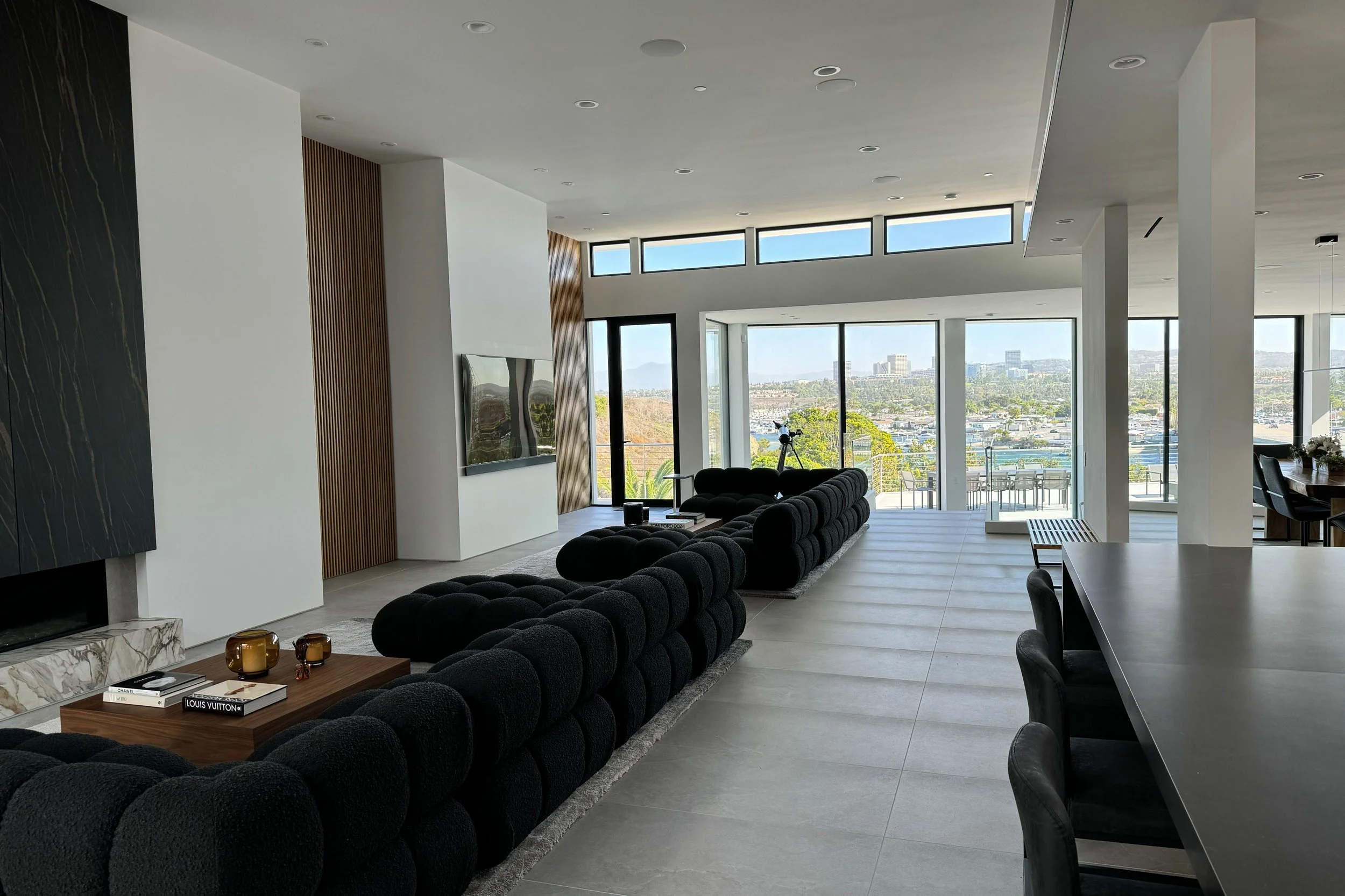 Modern living room with large windows showing cityscape, black sectional sofa, wooden coffee table with books and candles, and dining area with black chairs.