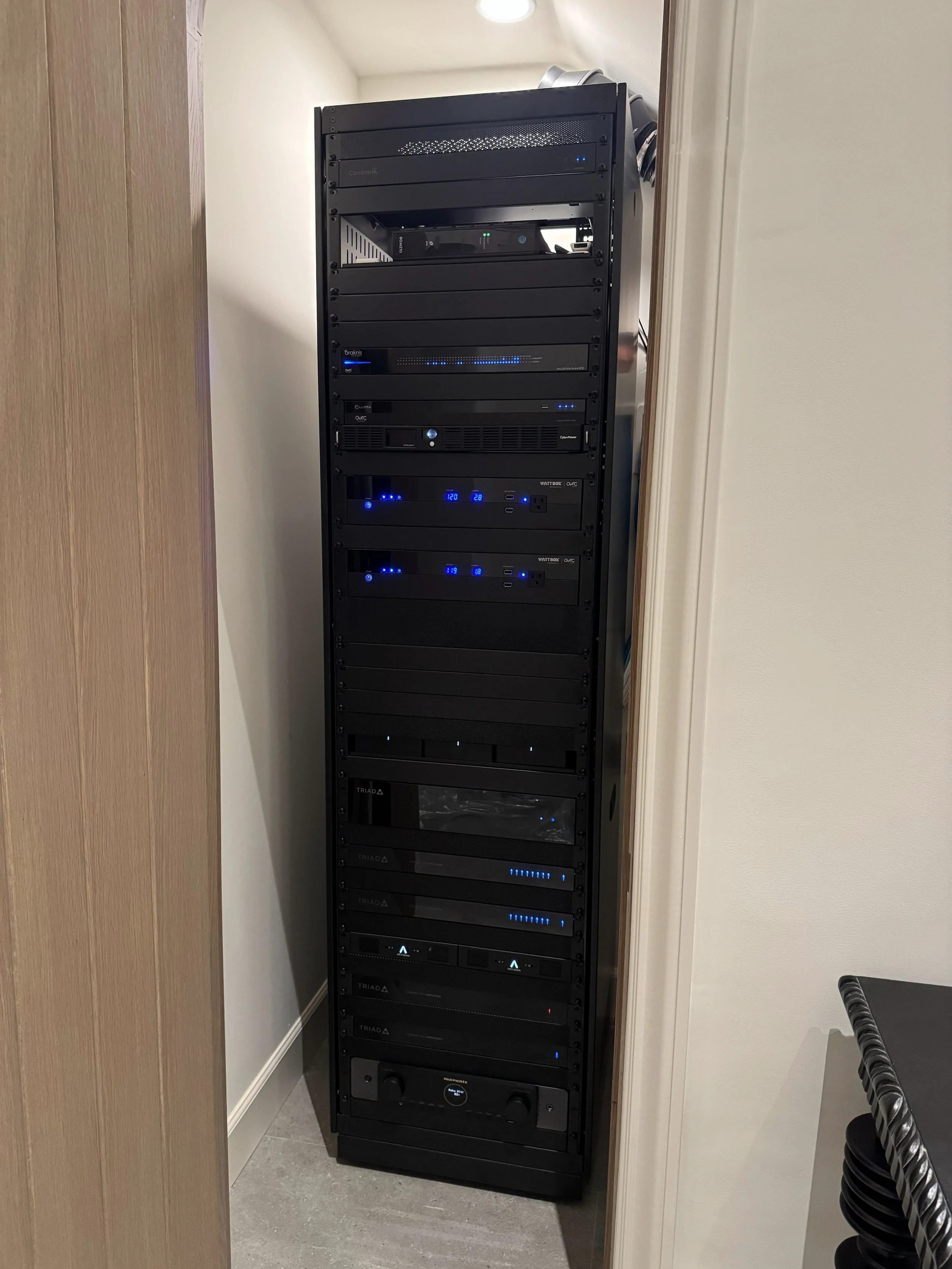 A tall black server rack with multiple electronic devices and blue LED lights, located in a small corner between a wooden door and white wall.