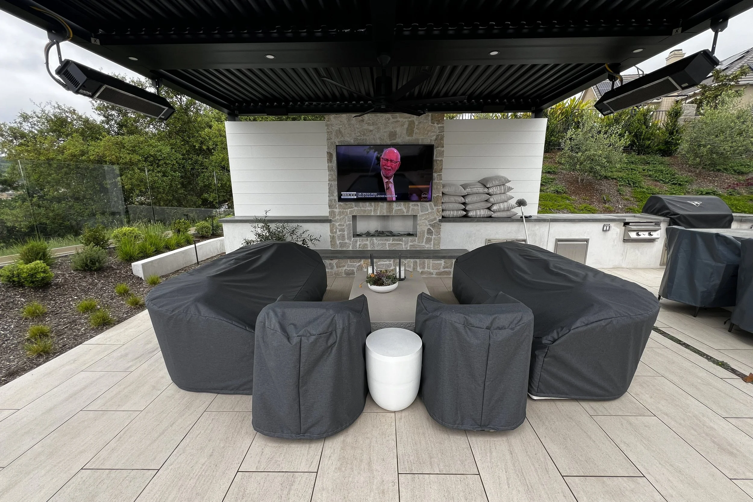 Outdoor patio with gray cushioned chairs, a small white side table, a stone fireplace with a TV, stacked pillows, and a grill, surrounded by green plants and trees.