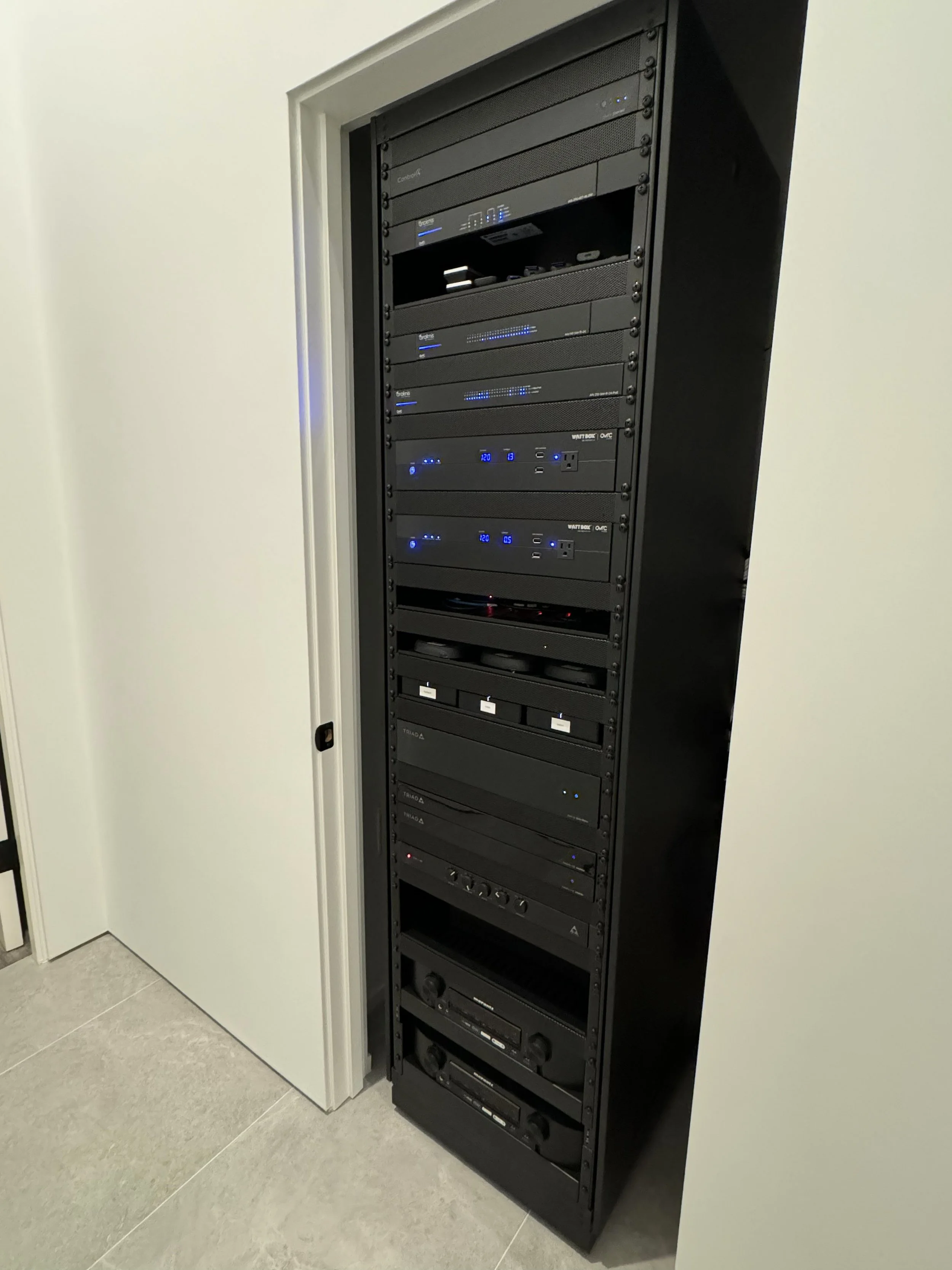 A black server rack with various electronic equipment and blue indicator lights is installed in a corner next to a white wall and door. The equipment includes switches, power supplies, and network devices.