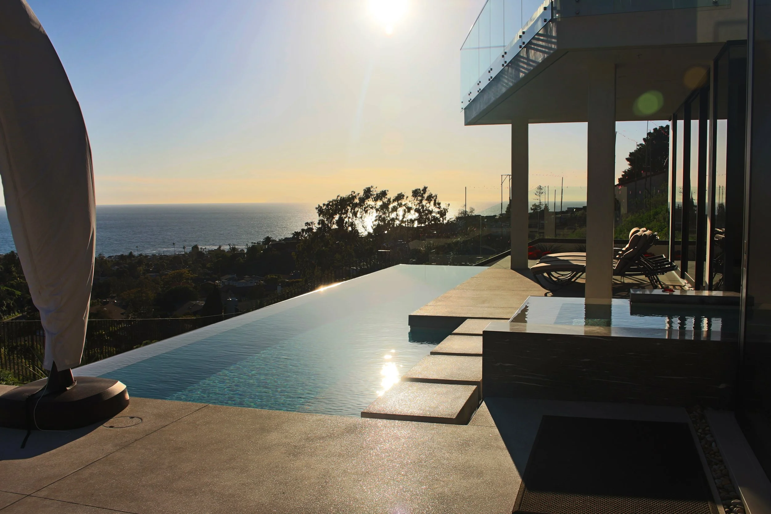 Modern home with a glass balcony, outdoor lounging chairs, and a view of the ocean in the background during sunset.
