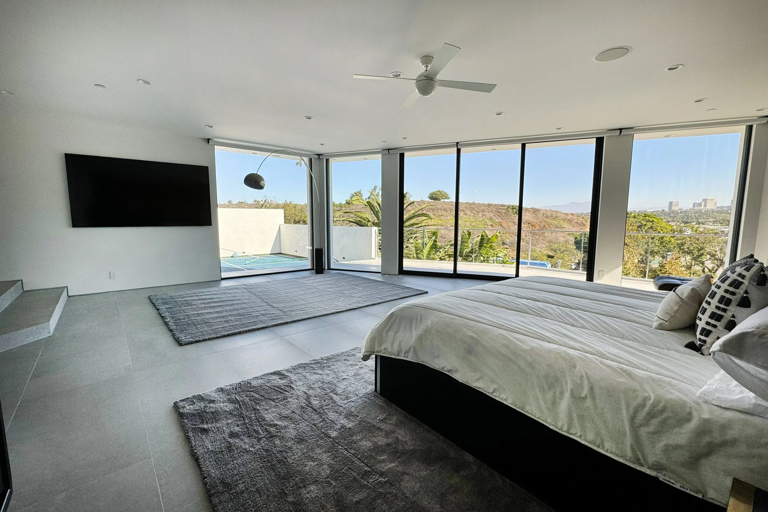Modern bedroom with large floor-to-ceiling windows overlooking trees and a distant cityscape, featuring a bed with white linens and decorative pillows, a wall-mounted television, a gray rug, a ceiling fan, and a sweeping natural light.
