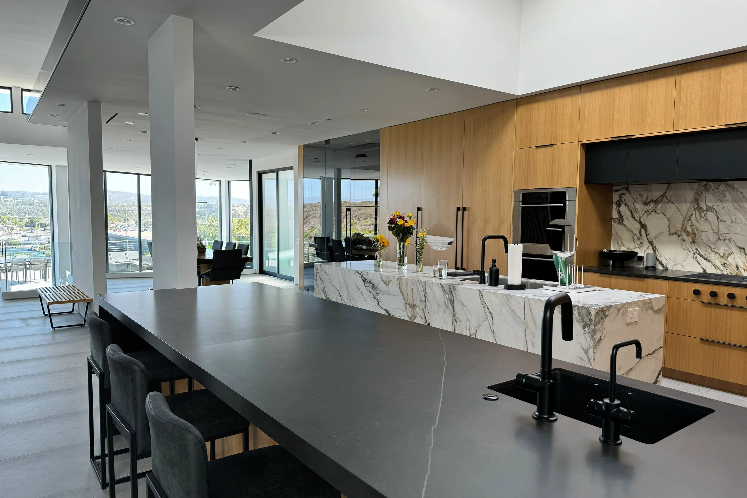 Modern kitchen and dining area with large windows, wooden cabinets, marble countertops, black sink, and bar seating.