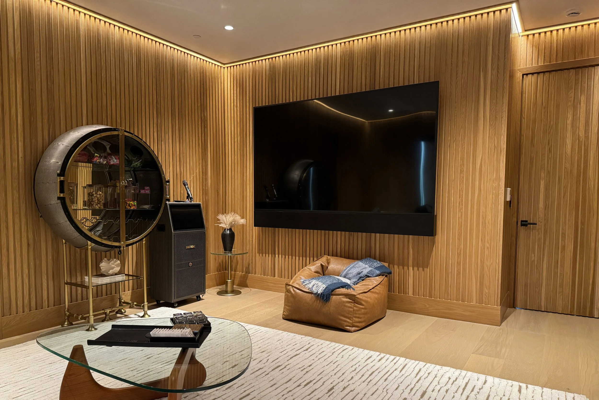 Interior of a room with wood-paneled walls, a large black flat-screen TV, a decorative circular shelf, a black speaker, a small side table with a vase, a leather ottoman with a blanket, a round glass coffee table, and a striped rug.
