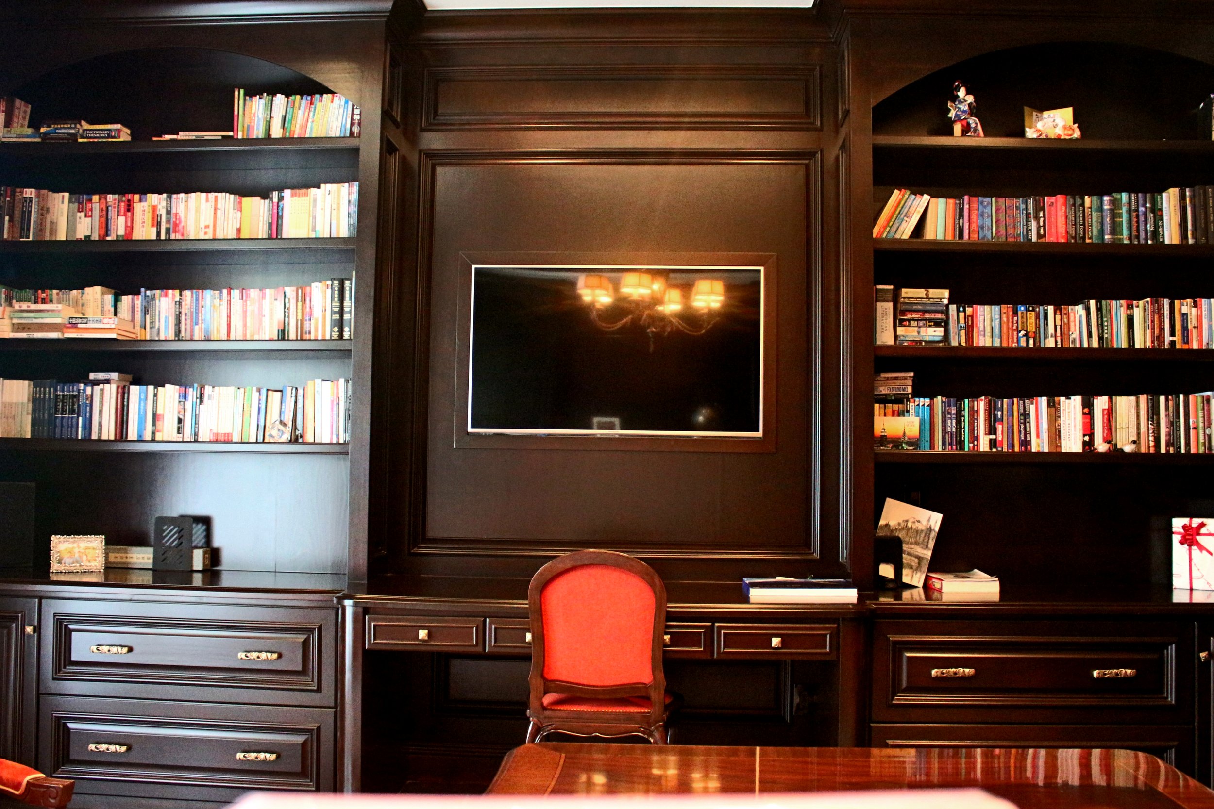 A dark wooden bookshelf unit with books and decorative items, mounted on a wall with a central mirror reflecting a chandelier, and a red-upholstered chair in front.