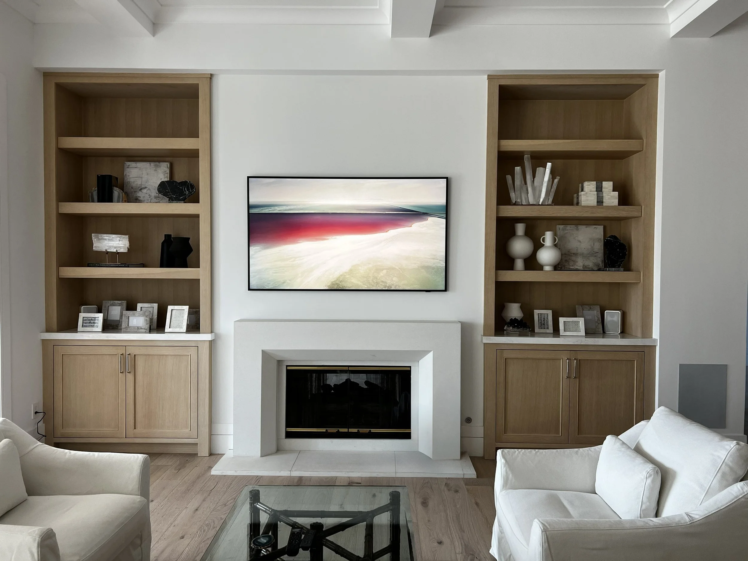 Living room with a white fireplace, two wooden bookshelves on either side, a television mounted above the fireplace displaying an abstract landscape, and seating including two beige armchairs. There is a glass coffee table with remote controls in the