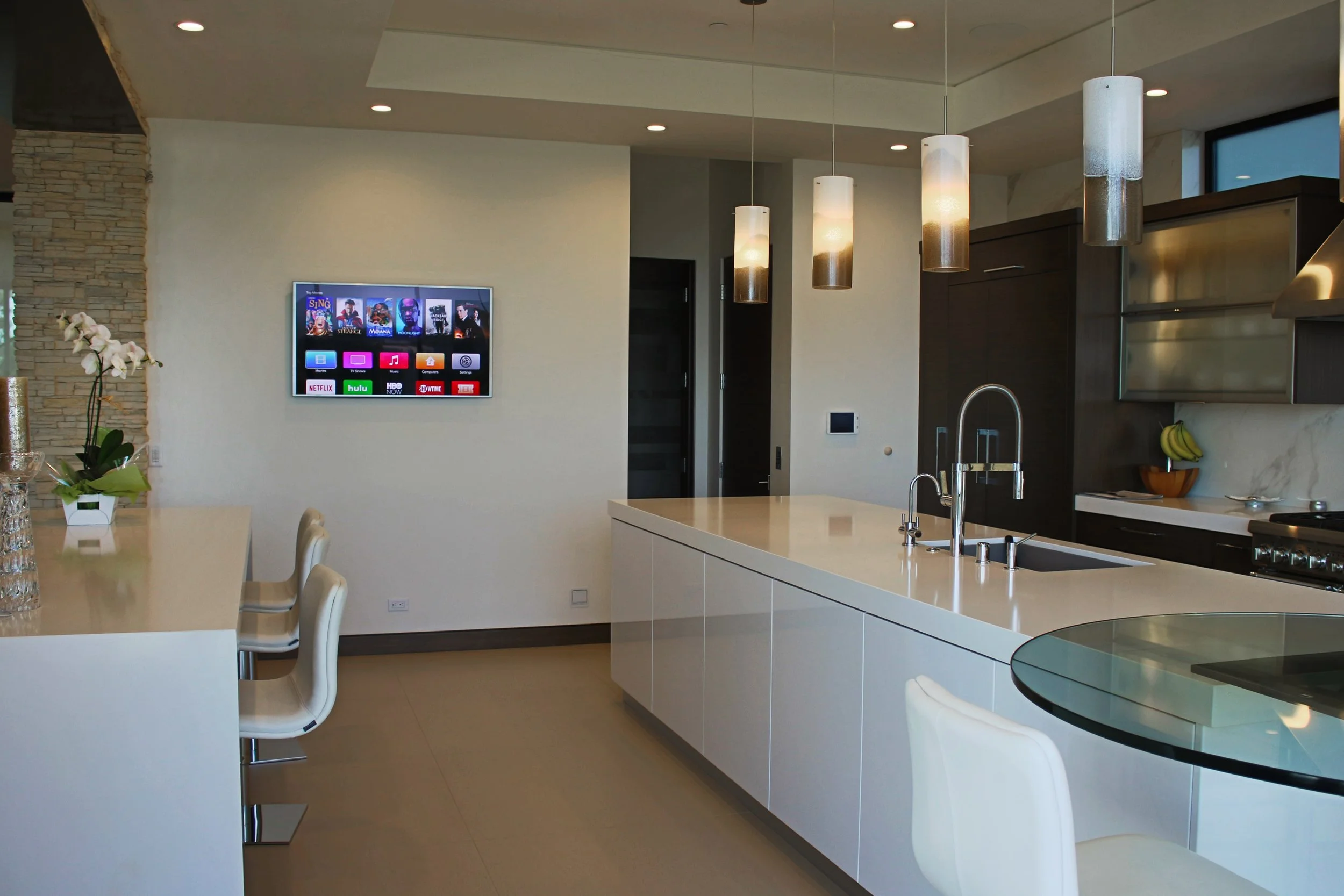 Modern kitchen with white island, dark cabinetry, pendant lights, a wall-mounted television, and a dining area with white chairs and a glass table.
