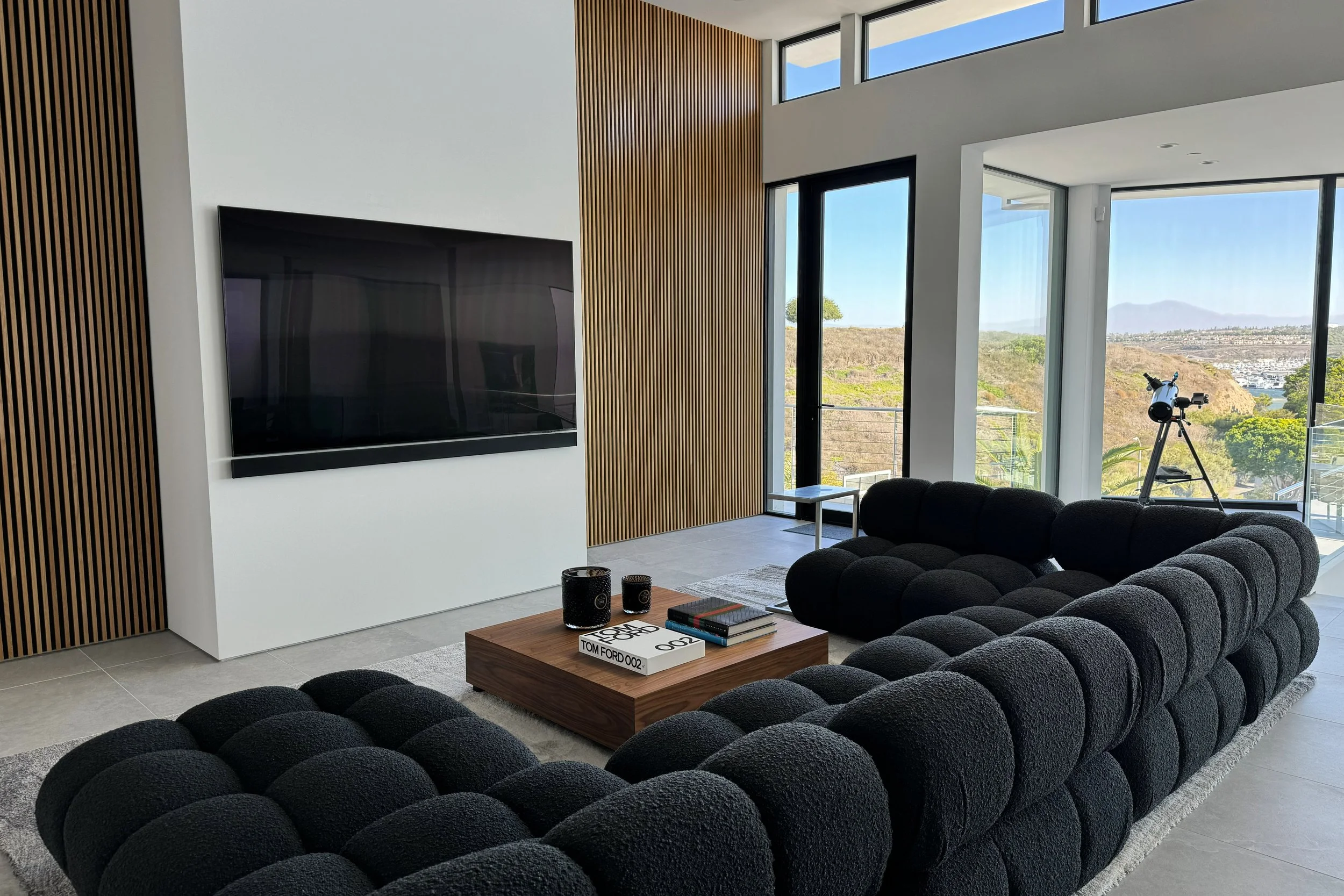 Modern living room with large windows showing a scenic outdoor view, a black sectional sofa, a wooden coffee table with books and decorative items, a flat-screen TV mounted on a white wall, a telescope near the window, and light-colored tiled floors 