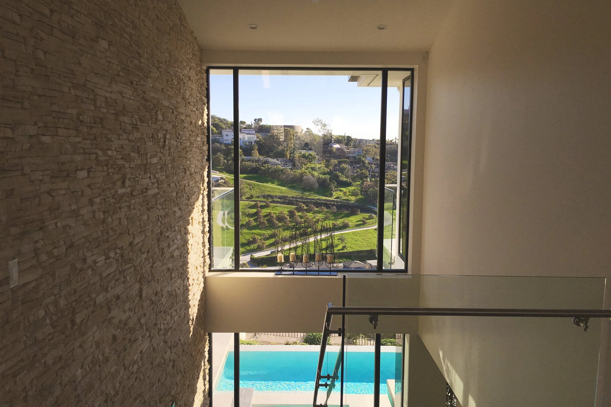 A city view through large floor-to-ceiling windows in a modern room, with a stone accent wall on the left and a white wall on the right. Visible are a swimming pool outside and greenery on a hillside with houses and trees in the background.