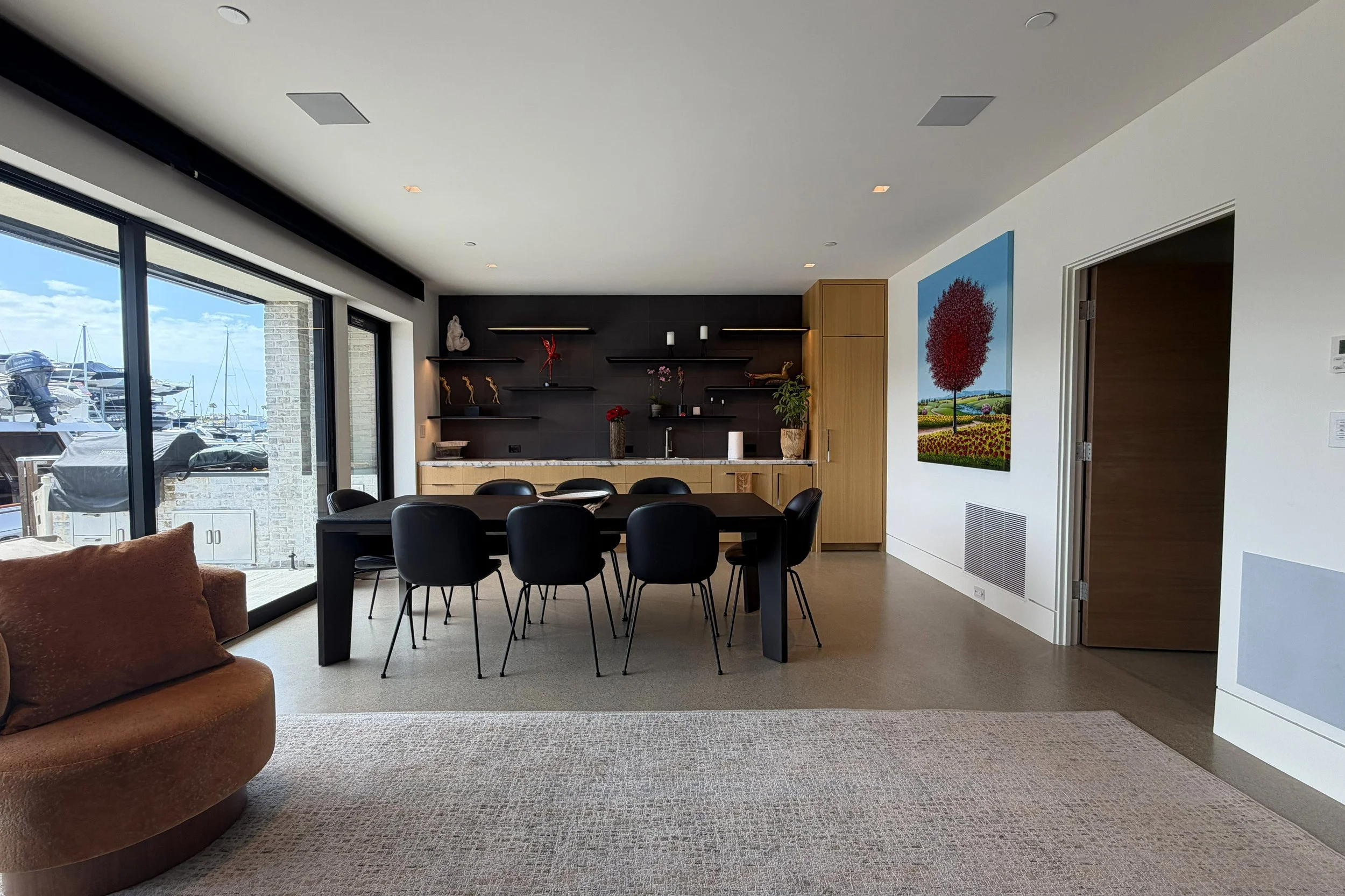 Modern living room with large glass doors opening to a marina with boats, contemporary black dining table with chairs, wall art of a colorful tree, and decorative items on black shelves.