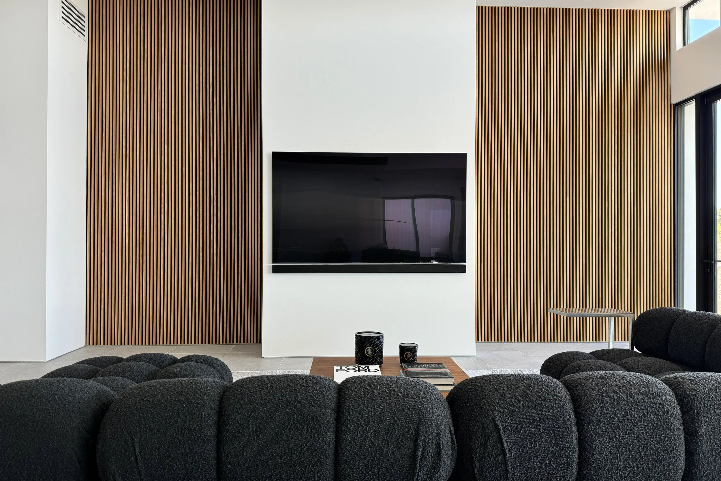 Modern interior with black sofas, a wooden slat wall, a wall-mounted TV, and a window with natural light.