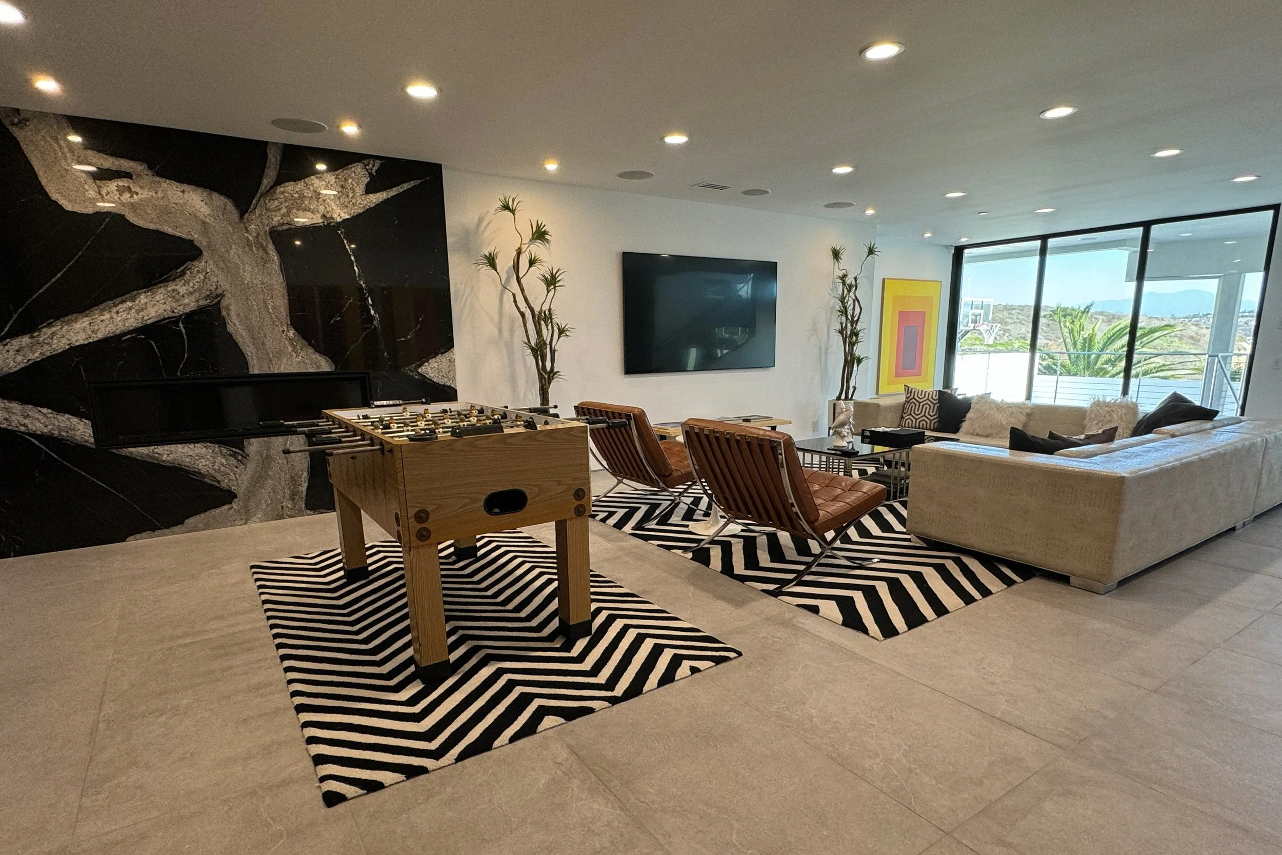 Modern living room with large glass windows showing outdoor landscape, featuring a black marble fireplace, potted plants, a flat-screen TV, art on the wall, and a seating area with a beige sectional sofa, wooden chairs, and decorative pillows, with a