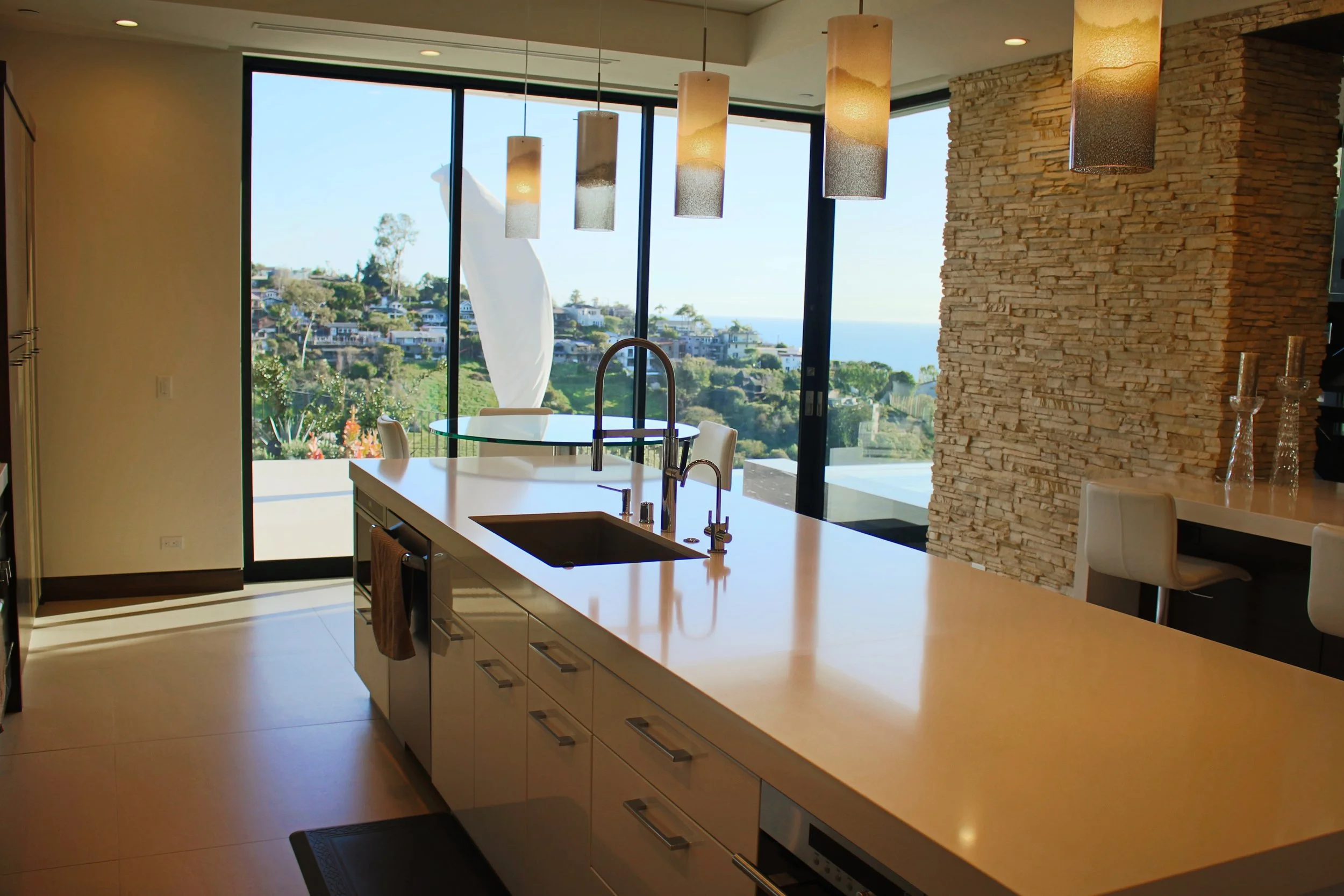 Modern kitchen with large windows showing a hillside view, pendant lights hanging above a island with a sink, and a textured stone wall.