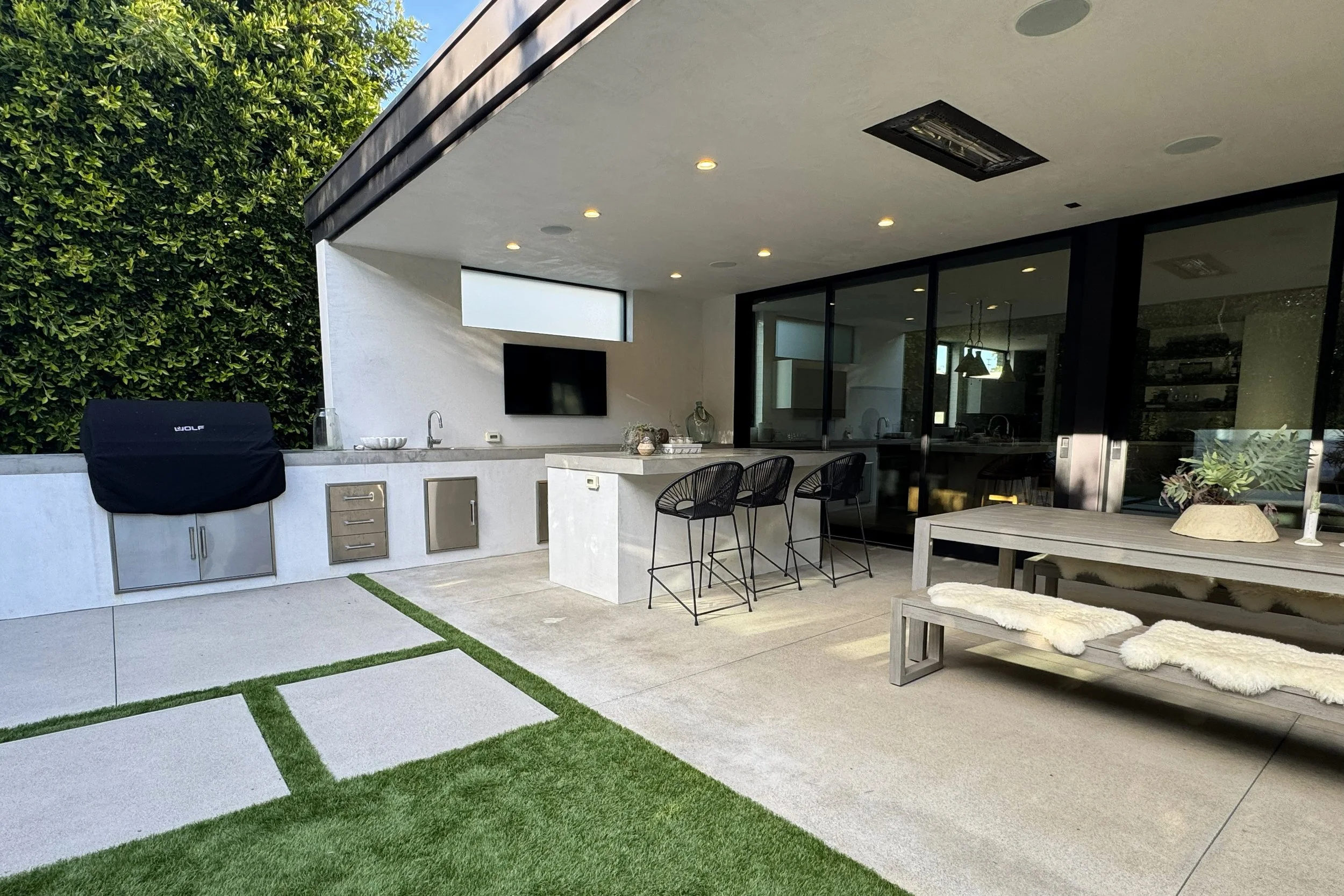 Modern outdoor patio area with a built-in grill, black chairs around a table, a mounted TV, and a view into an indoor space through large glass sliding doors. The patio features concrete and artificial grass flooring, with lush green hedges on one si