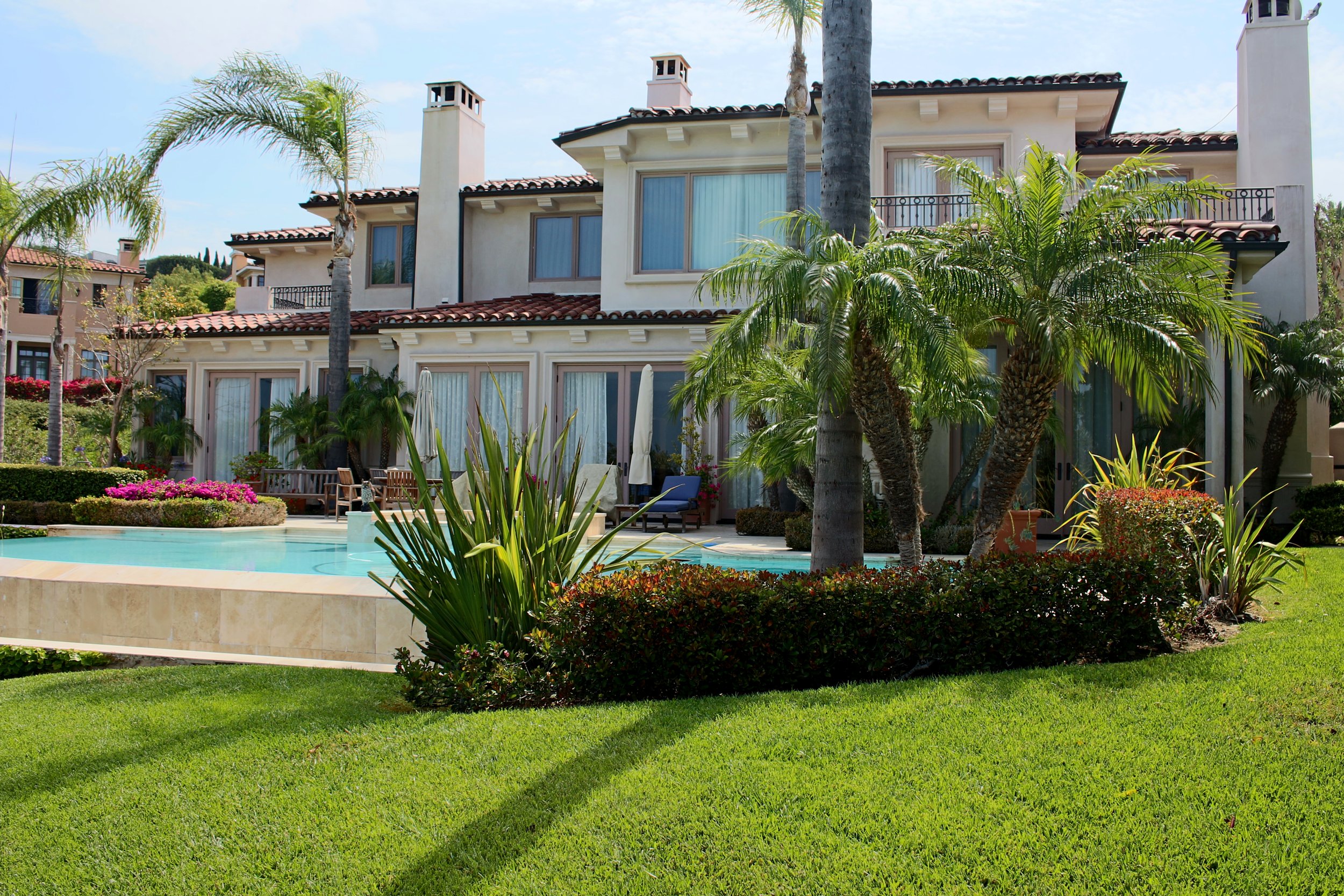 Luxury white residential building with a red tile roof, surrounded by palm trees, lush green lawn, and a swimming pool in the foreground under a partly cloudy sky.