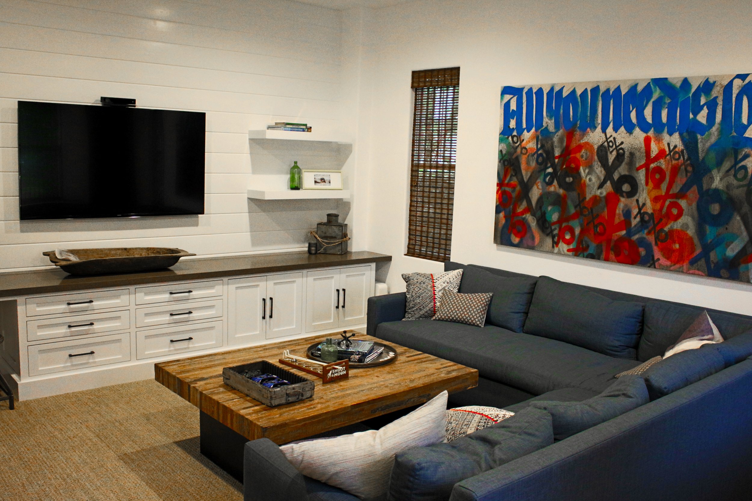 Living room with white shiplap walls, a mounted flat-screen TV, a corner window with bamboo shade, abstract art painting, gray sectional sofa with patterned pillows, wooden coffee table with decor items, and a built-in white cabinet with drawers.