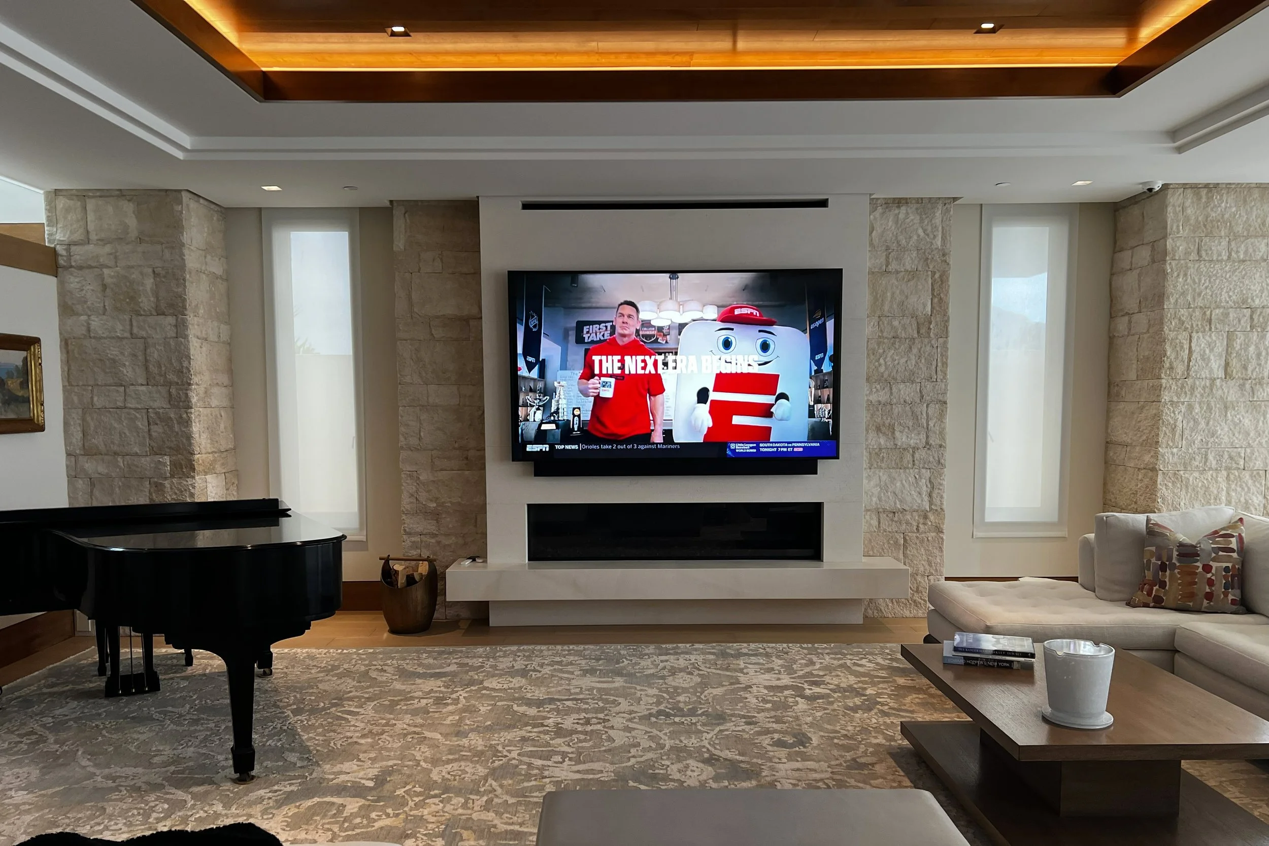 Living room with a mounted flat-screen TV showing a sports news broadcast, a black grand piano on the left, a plush beige sofa on the right with colorful patterned pillows, a coffee table with books and a white vase, and a large beige and gray patter
