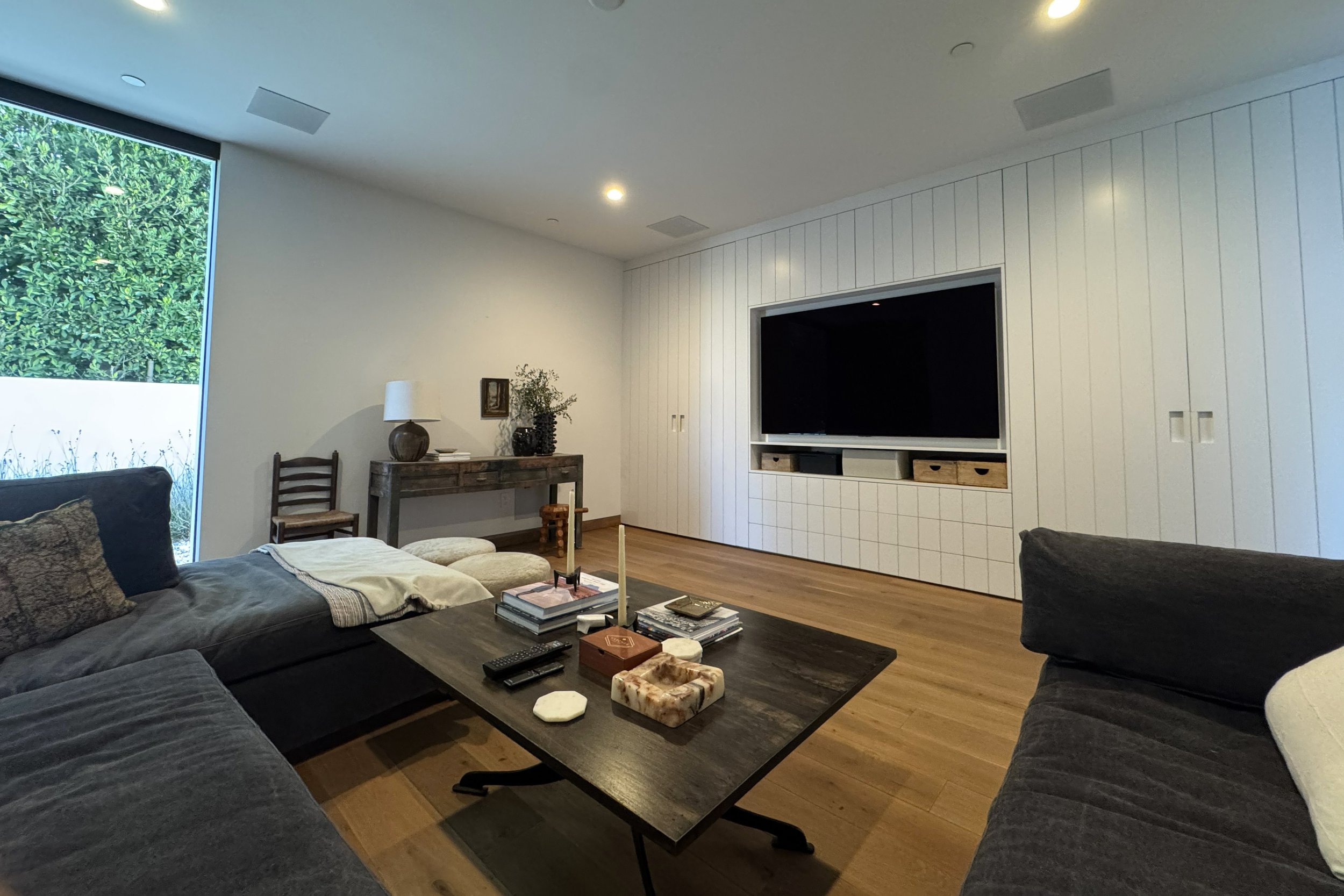 Living room with dark gray sofas, a wooden coffee table with books and decorative items, a wooden console table with a table lamp and plants, a flat-screen TV, and large window with greenery outside.