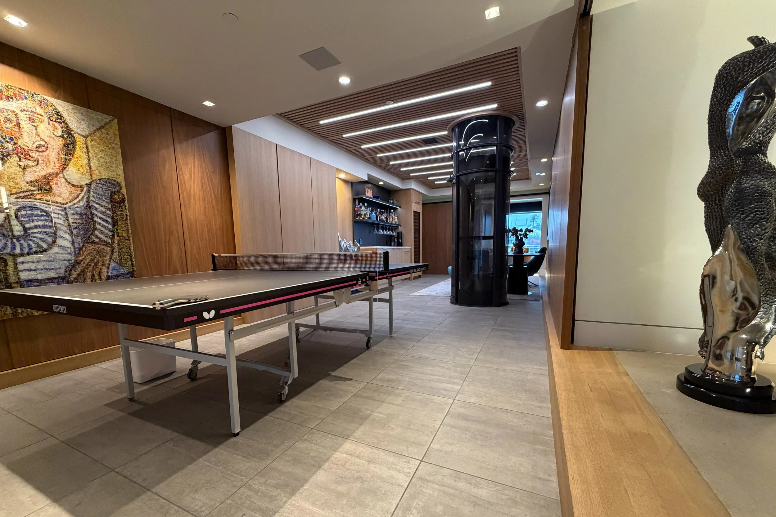 An indoor recreation room with a ping pong table, a bar area with shelves of liquor, a glass elevator, and decorative art sculptures.