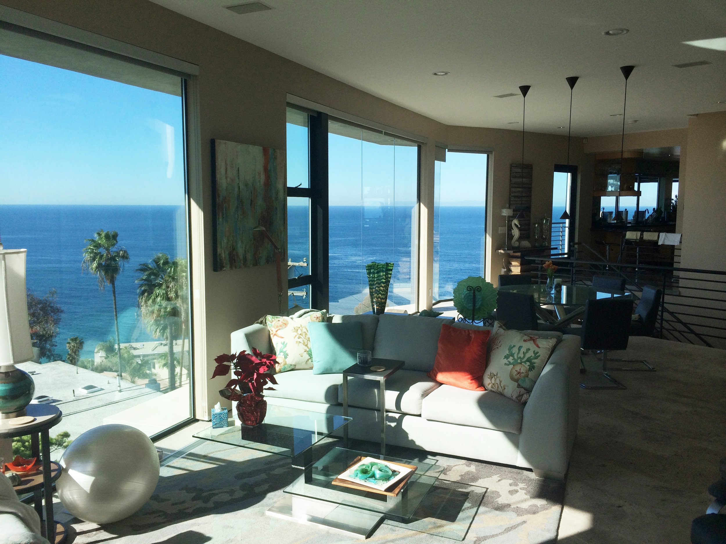 Living room with large floor-to-ceiling windows showing ocean view, white sofa with colorful pillows, glass coffee table with decorative items, plants, and modern furniture.