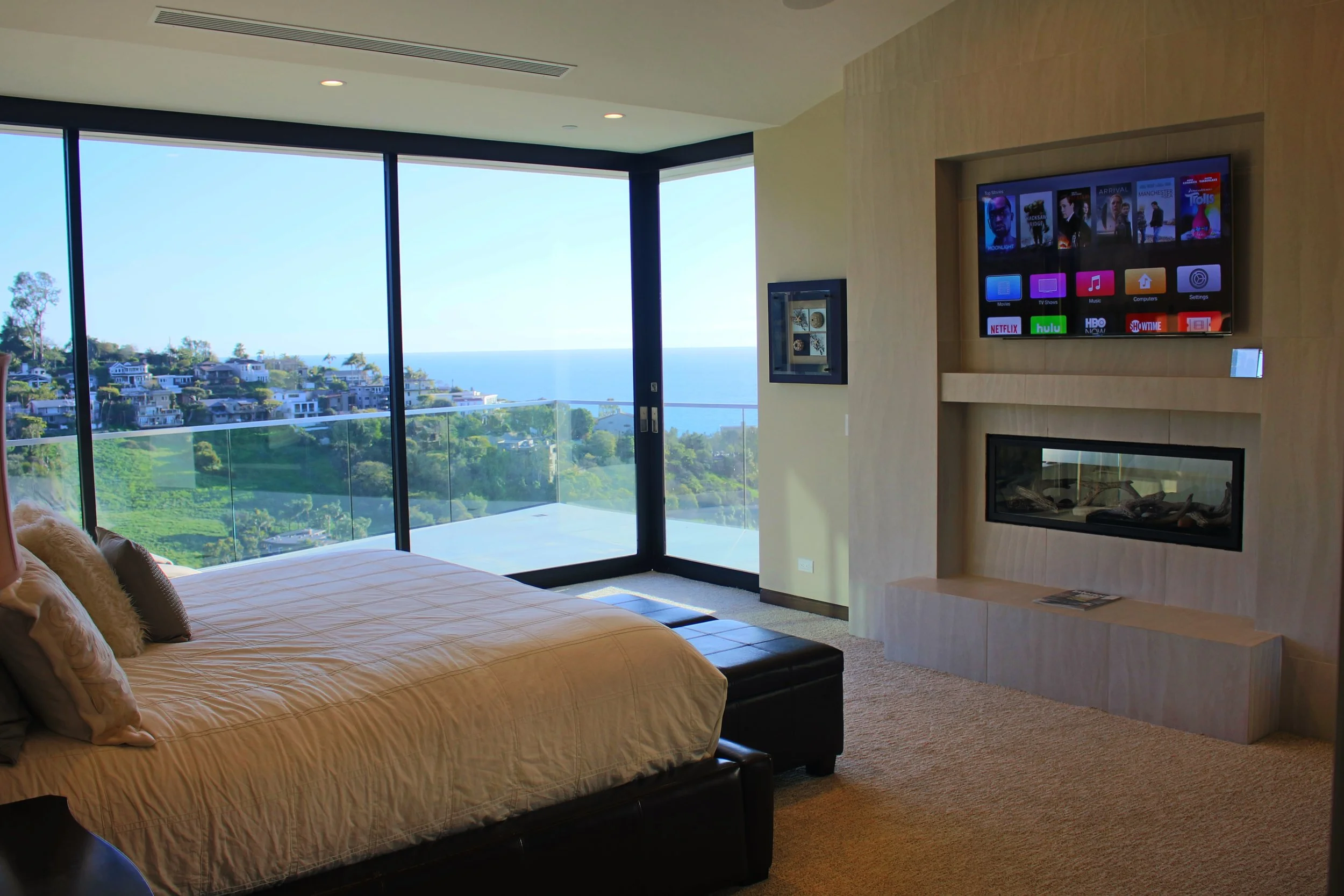 A bedroom with a large glass window and a view of a hilly neighborhood with trees, overlooking the ocean. The bed has beige bedding and pillows, and there is a wall-mounted television to the right of the window.