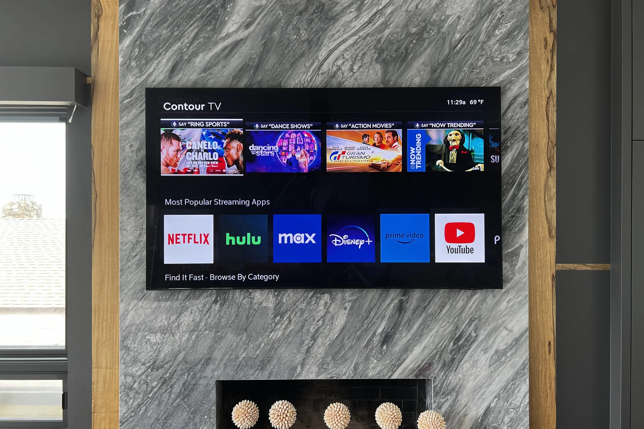 A flat-screen television mounted on a gray marble wall surrounded by a wooden frame. The TV displays streaming app icons and categories, with the current time 11:29 am and temperature 69°F at the top right corner. Part of a window with closed blinds 