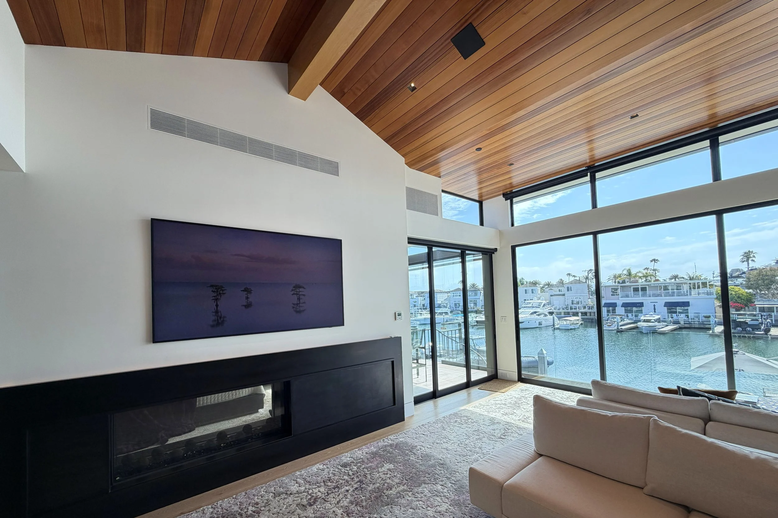 Modern living room with large windows overlooking a marina with boats, wood-paneled ceiling, white walls, and a wall-mounted TV above a black fireplace.