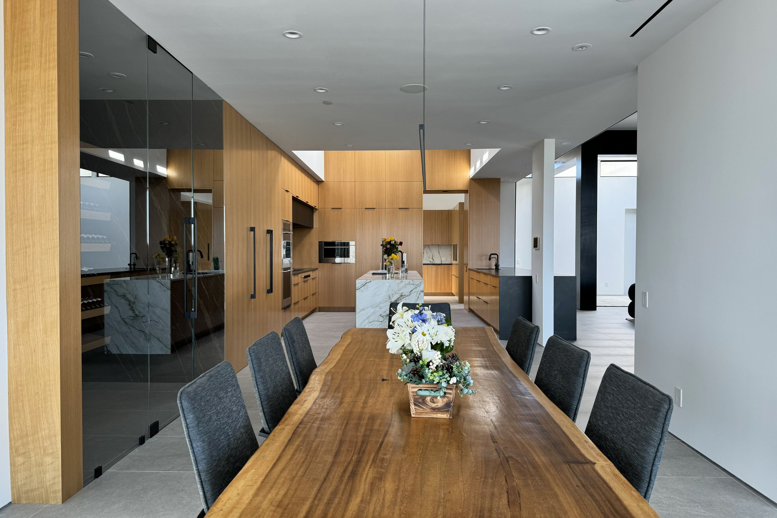 Modern kitchen and dining area with wooden cabinets, marble island, glass sliding door, and a dining table with floral centerpiece.