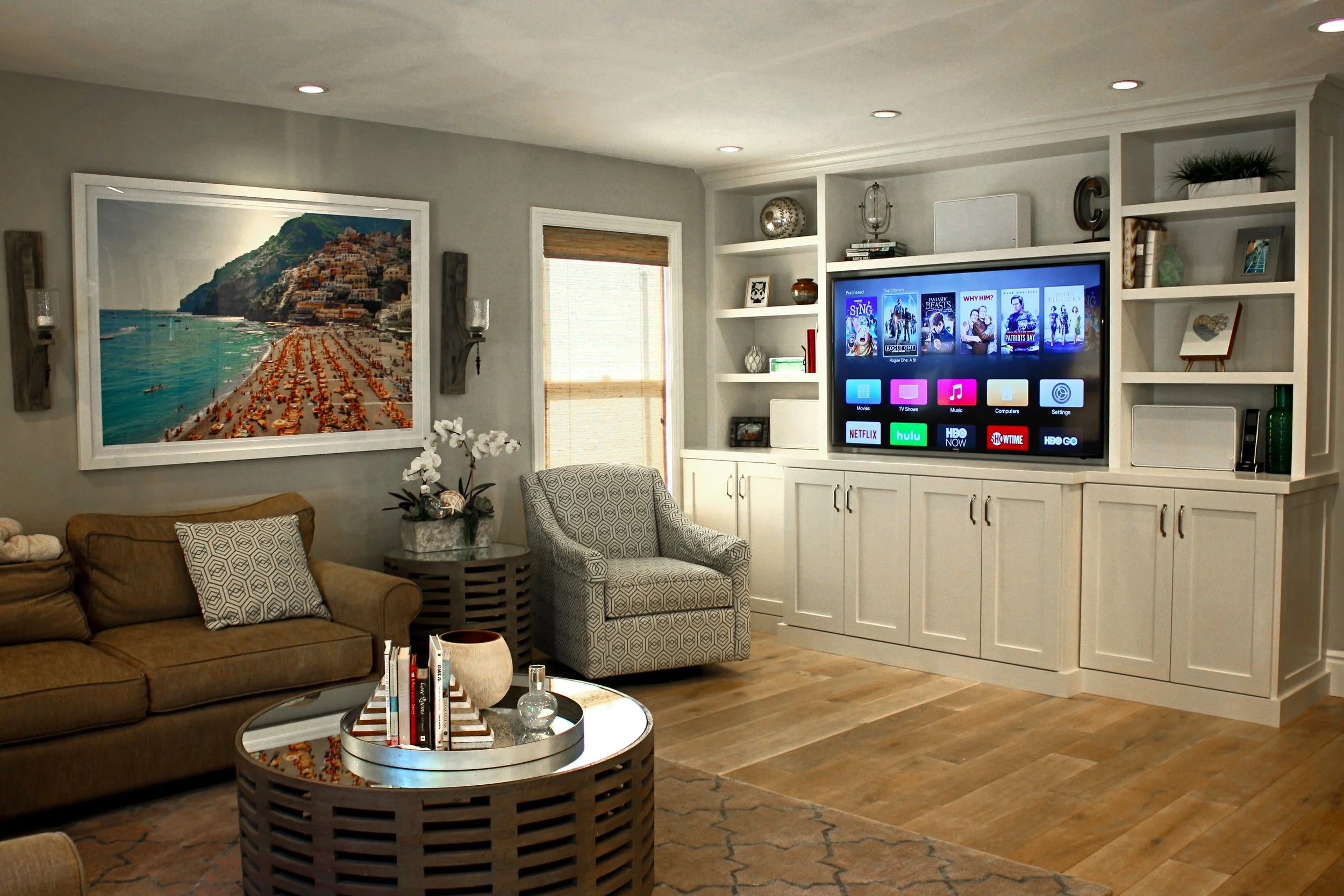 A living room with a brown sofa and a glass coffee table holding books and a decorative vase. There are patterned armchairs and a large framed photo of a beach scene on the wall. The entertainment center has a television displaying streaming service 