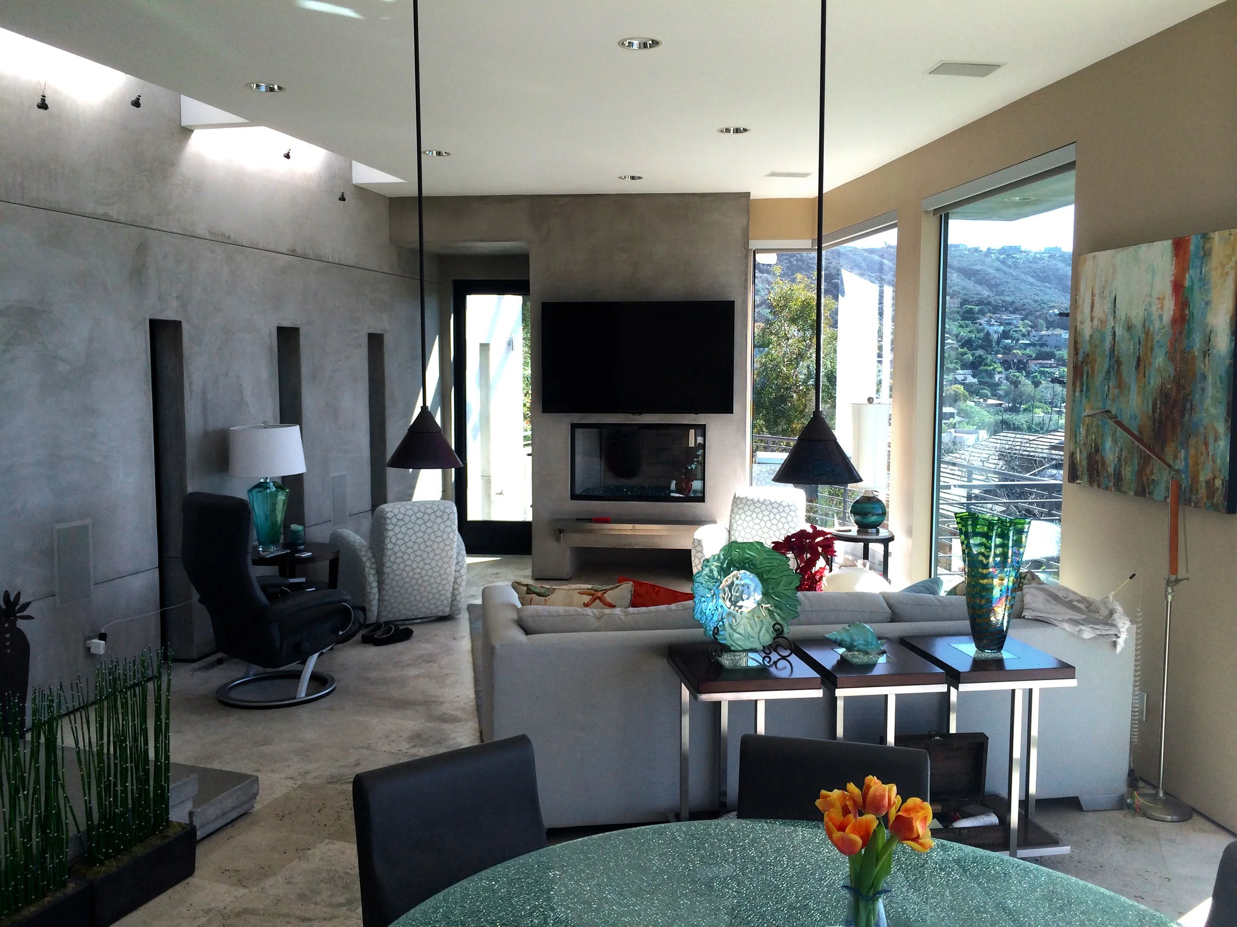 Modern living room with large windows showing a hillside view, featuring a flat-screen TV, fireplace, artwork, and various decorative glass vases on tables, with seating and plants.