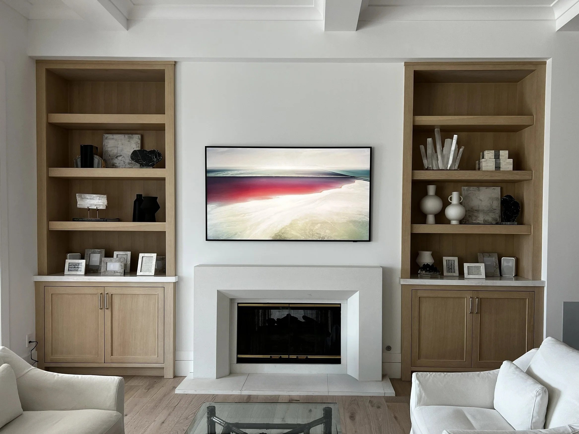 Living room with a fireplace flanked by built-in wooden bookshelves, a mounted flat-screen TV above the fireplace, and white upholstered chairs in the foreground.