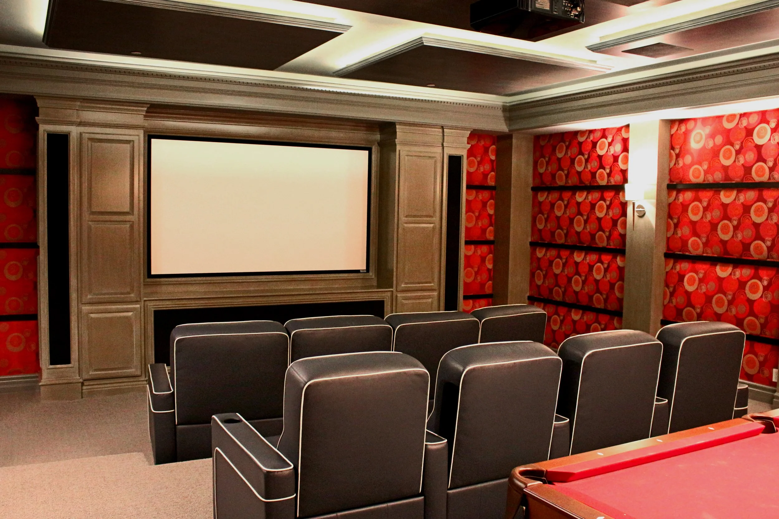 An empty movie theater or screening room with rows of black cushioned seats, a large projection screen, and red patterned wallpaper on the walls.
