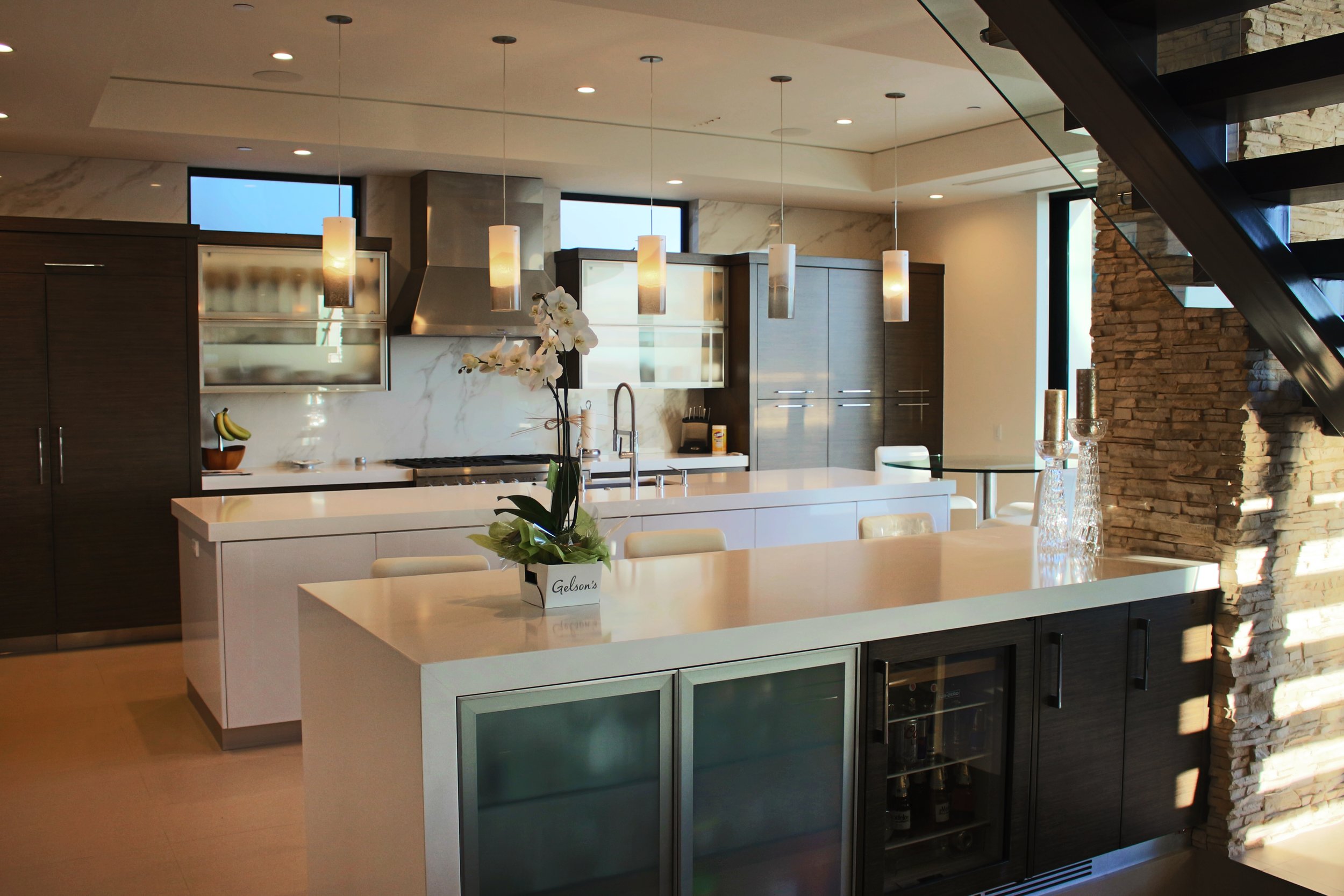 Modern kitchen with white island counter, dark wood cabinets, stone wall accent, hanging pendant lights, and orchids in a vase on the counter.