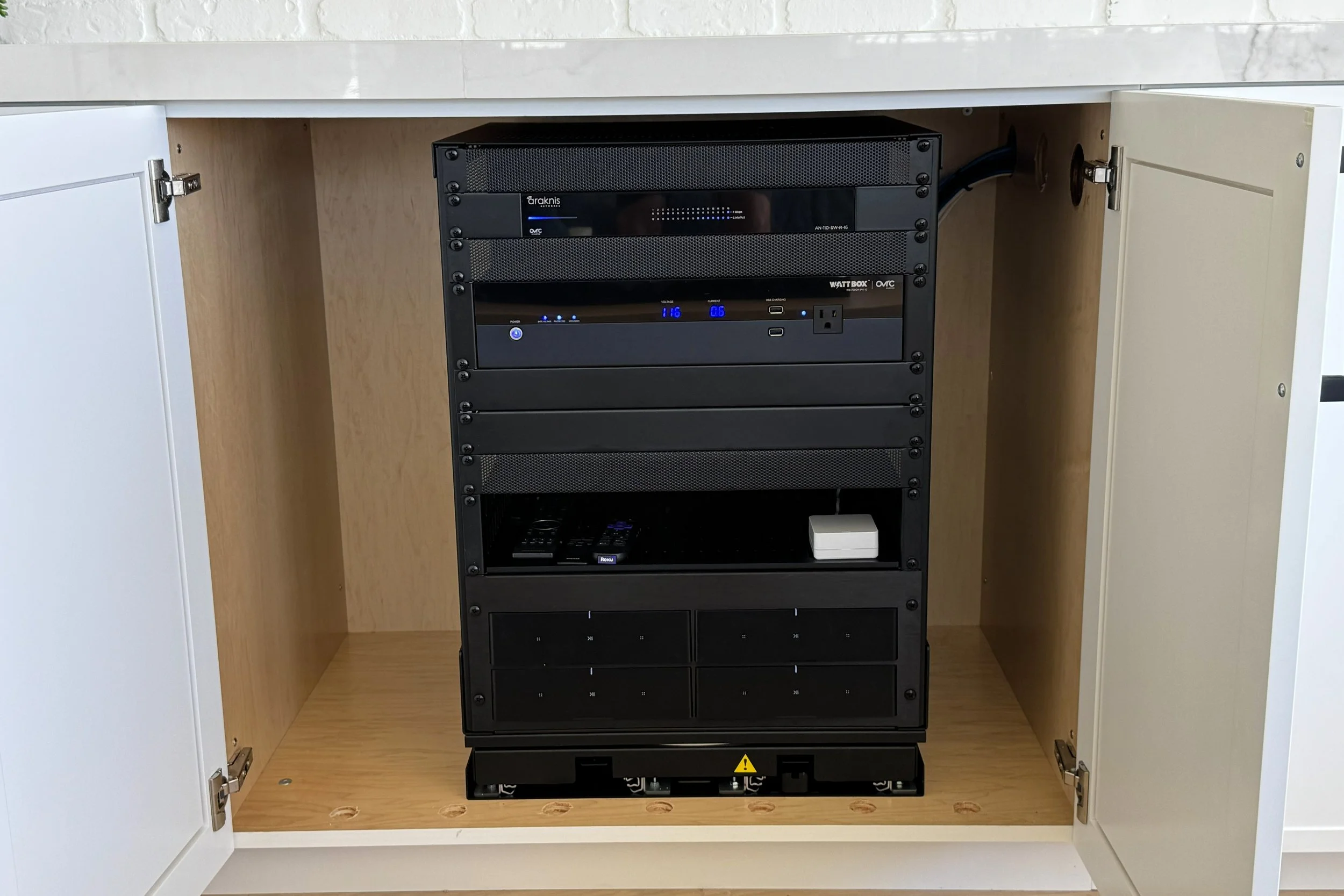 Inside a cabinet with its doors open, revealing electronic equipment stacked on a black rack, including a server, a power strip, and other devices, with a wooden interior and white exterior doors.